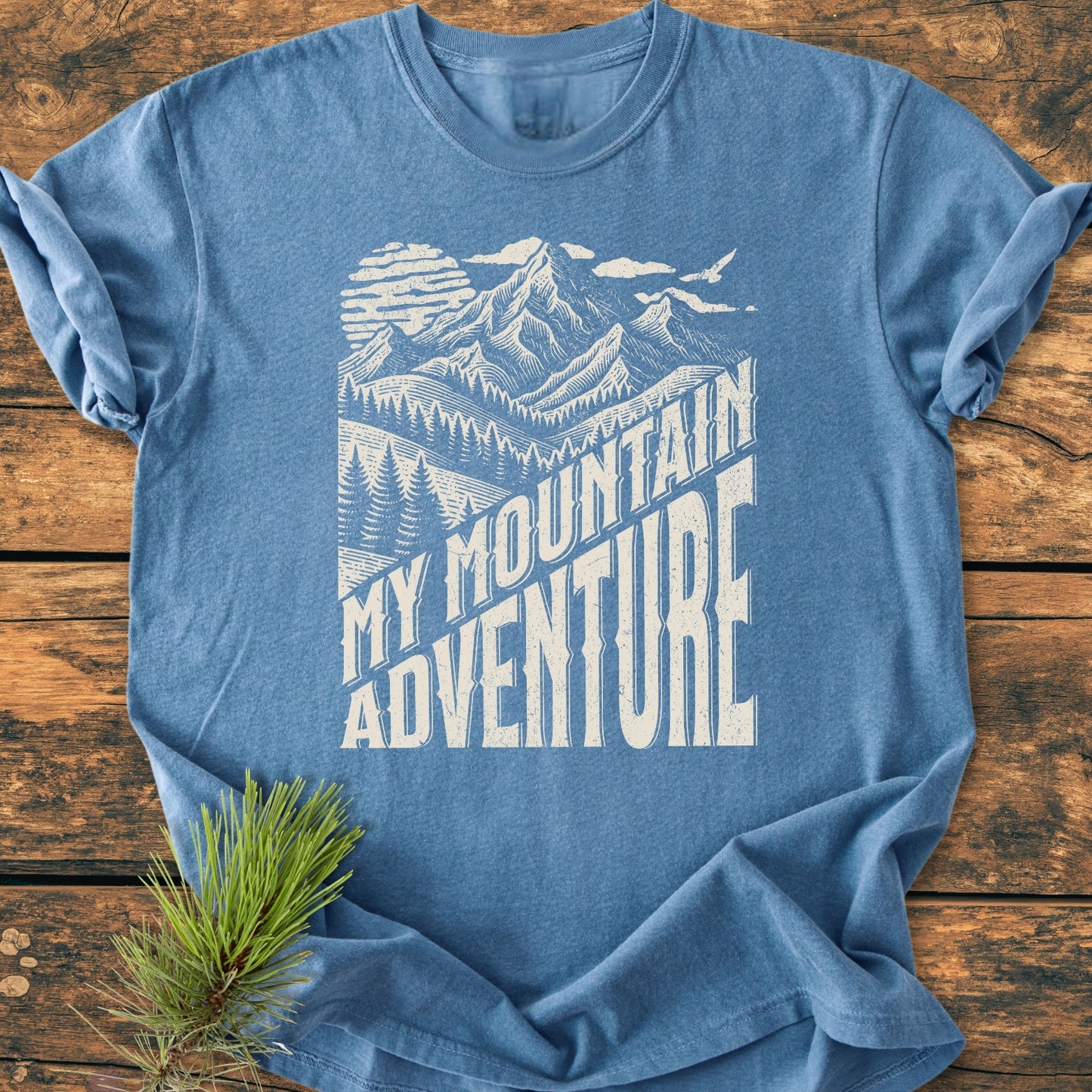 My Mountain Adventure - Vintage Wash Tee featuring a stylized mountain scene with text, displayed on a wooden surface. A pine needle sprig adds a natural touch, highlighting the shirt's outdoor theme.