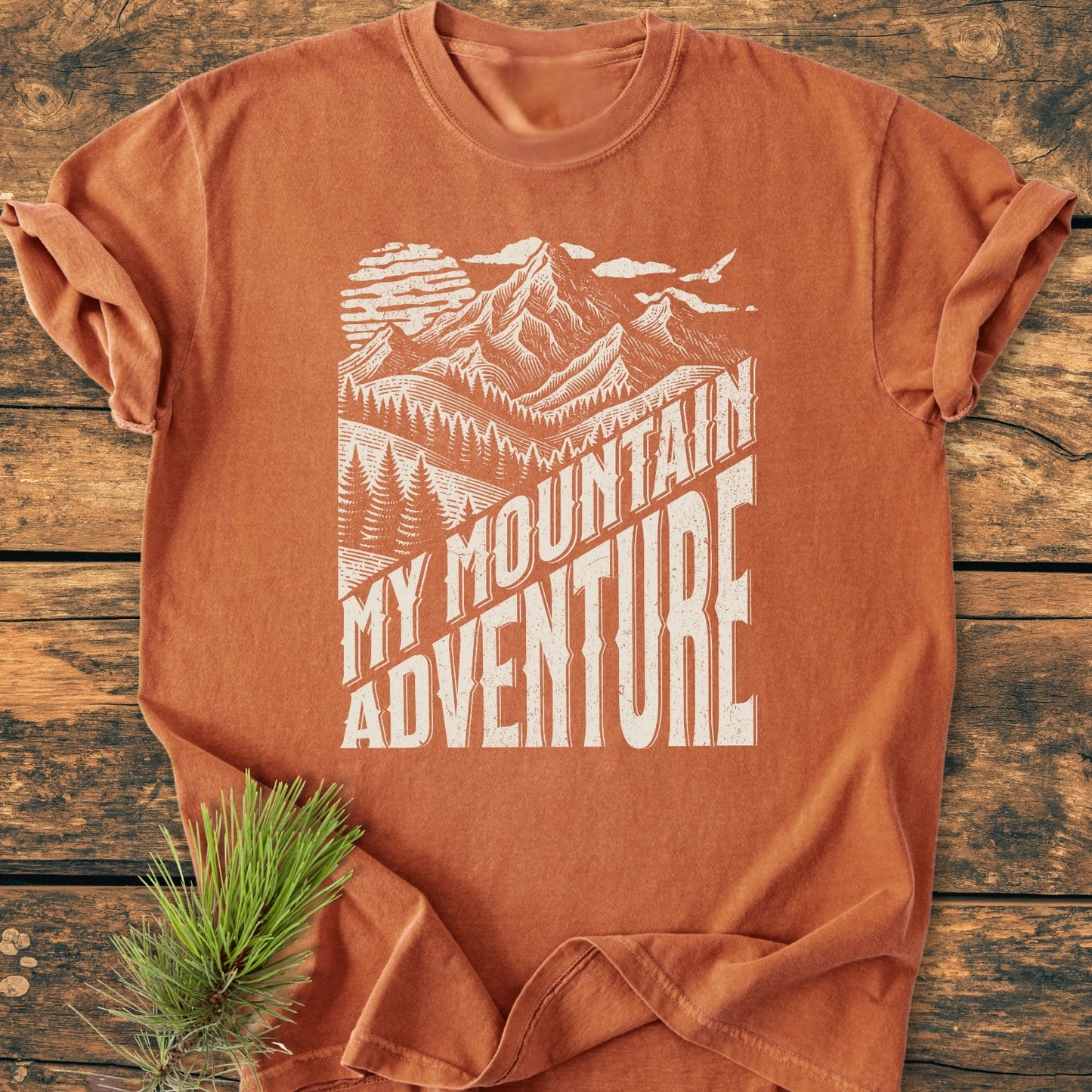 My Mountain Adventure - Vintage Wash Tee features a burnt-orange hue with a white mountain landscape print. Laid on a wooden surface, it highlights the tee's relaxed fit and soft-washed fabric.