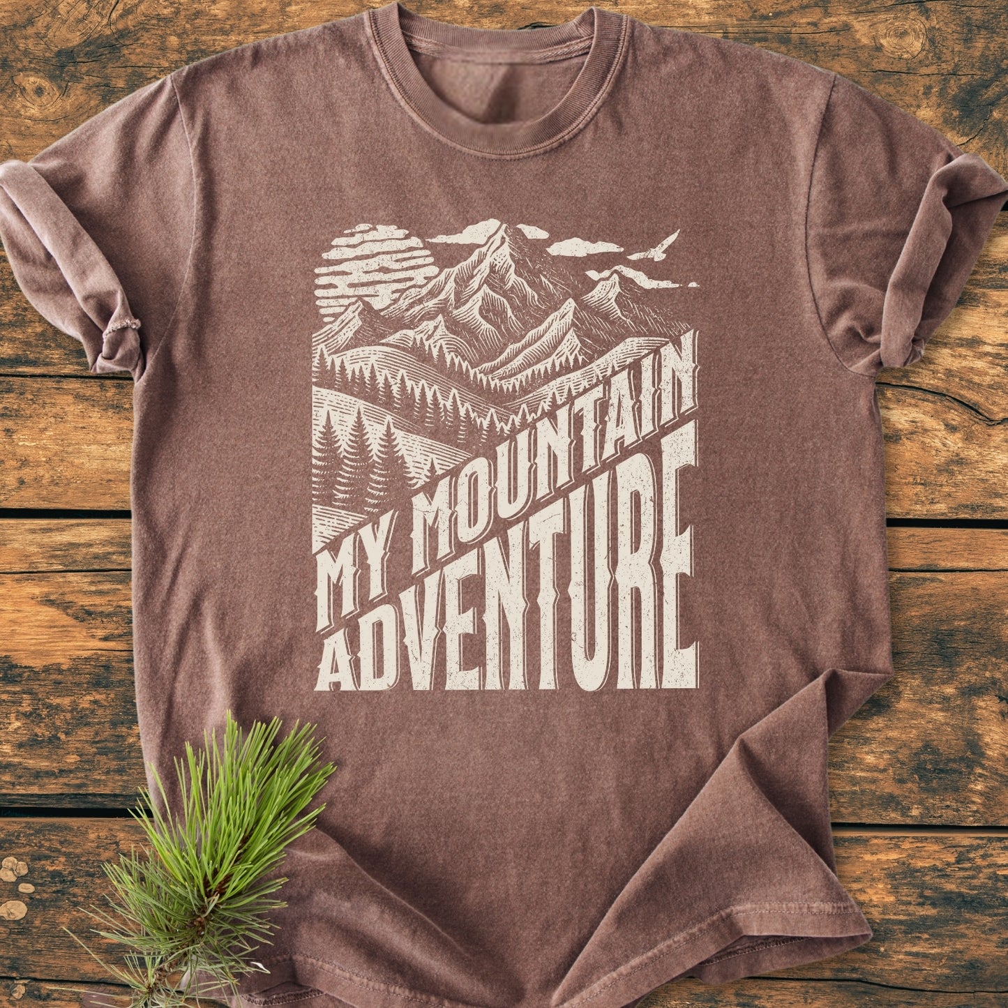 My Mountain Adventure - Vintage Wash Tee features a block-print mountain and forest graphic with a sun and bird. Laid flat, it emphasizes its relaxed fit and durable, soft-washed cotton fabric.