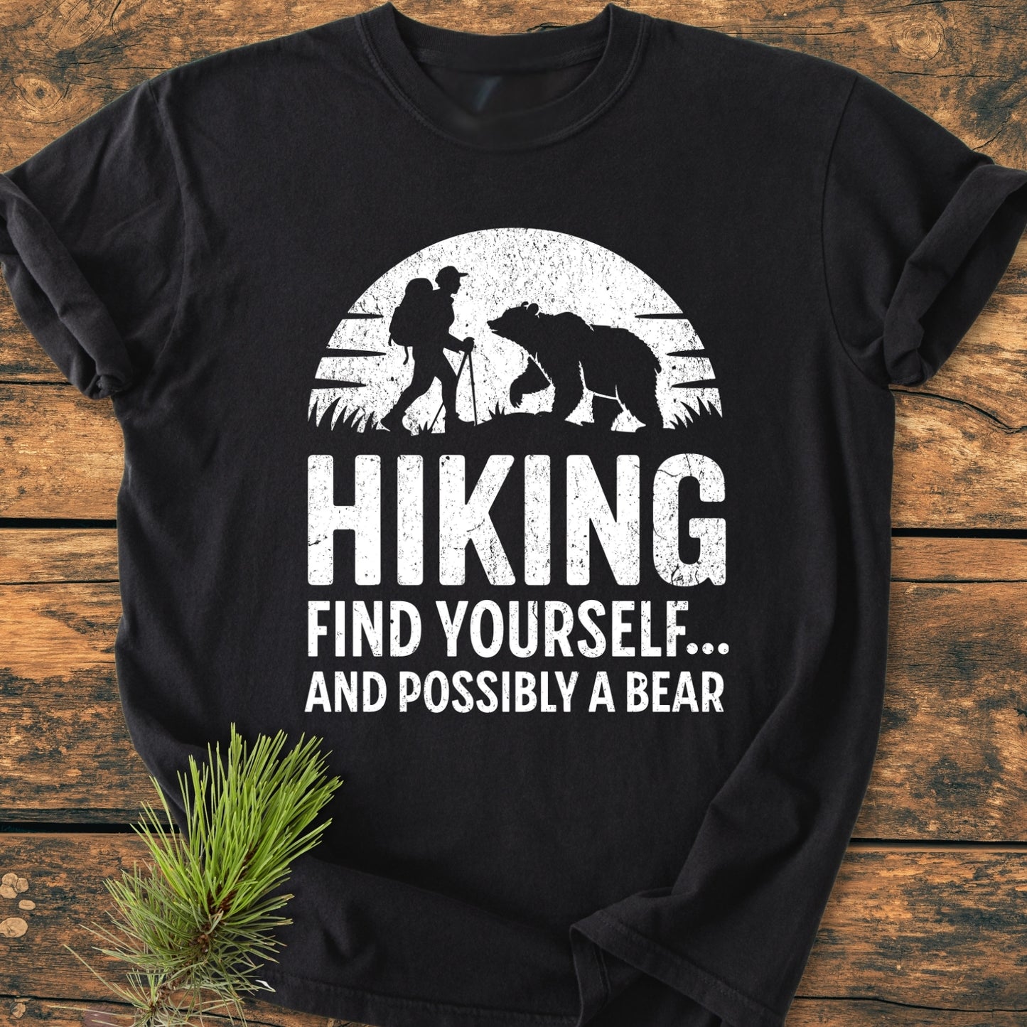 Find Yourself And Possibly A Bear - Vintage Wash Tee features a distressed graphic of a hiker and bear beneath a sun/moon on a black T-shirt, laid on a wooden surface.