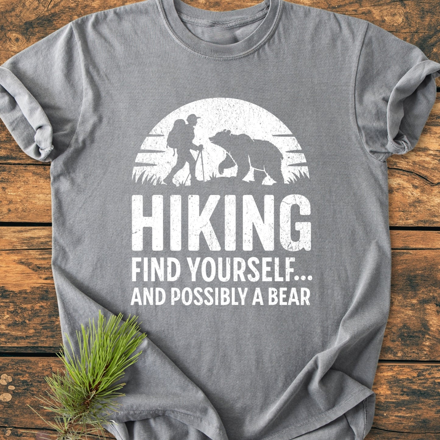 Find Yourself And Possibly A Bear - Vintage Wash Tee features a white backpacker-bear graphic on heather-gray fabric, displayed on a wooden surface with a pine sprig, highlighting its outdoor theme.