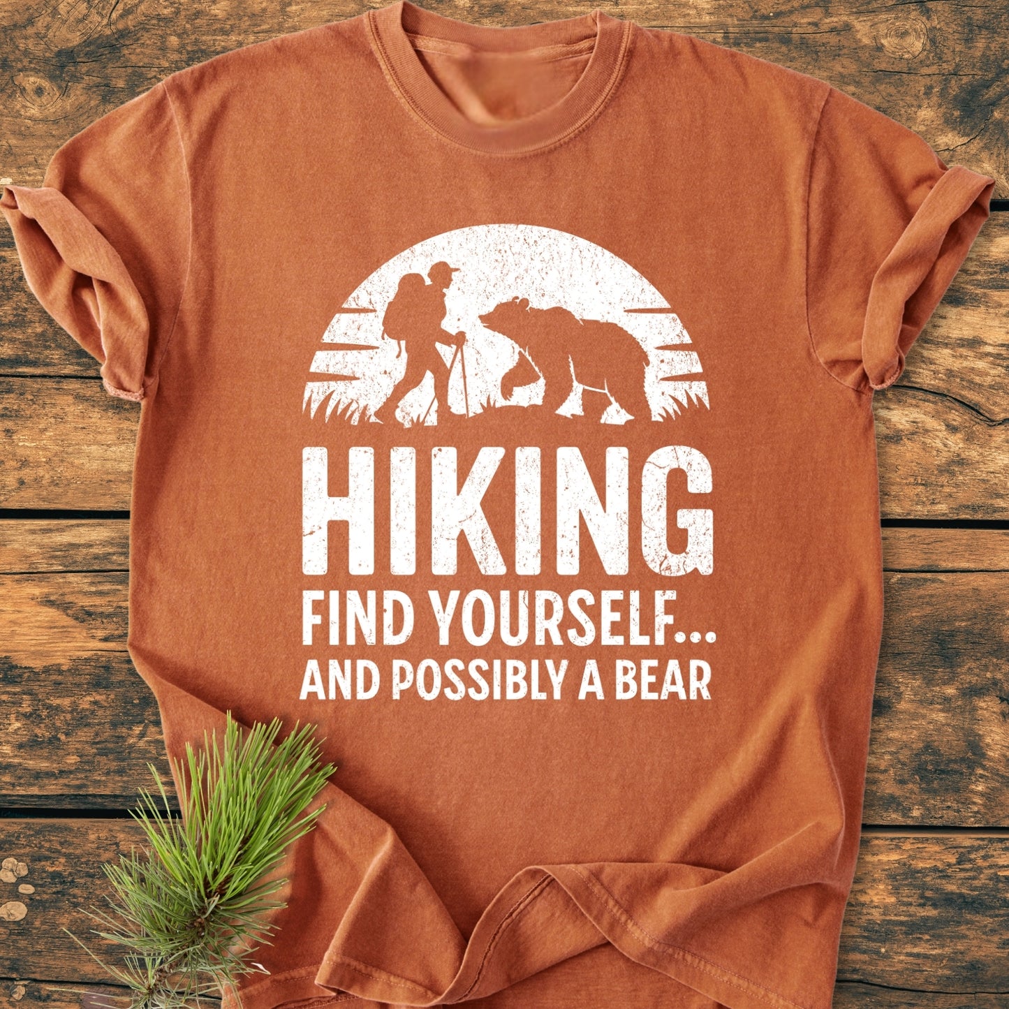 Find Yourself And Possibly A Bear - Vintage Wash Tee features a stylized hiker and bear graphic on a rust-orange T-shirt, laid flat with a pine sprig, capturing outdoor adventure spirit.