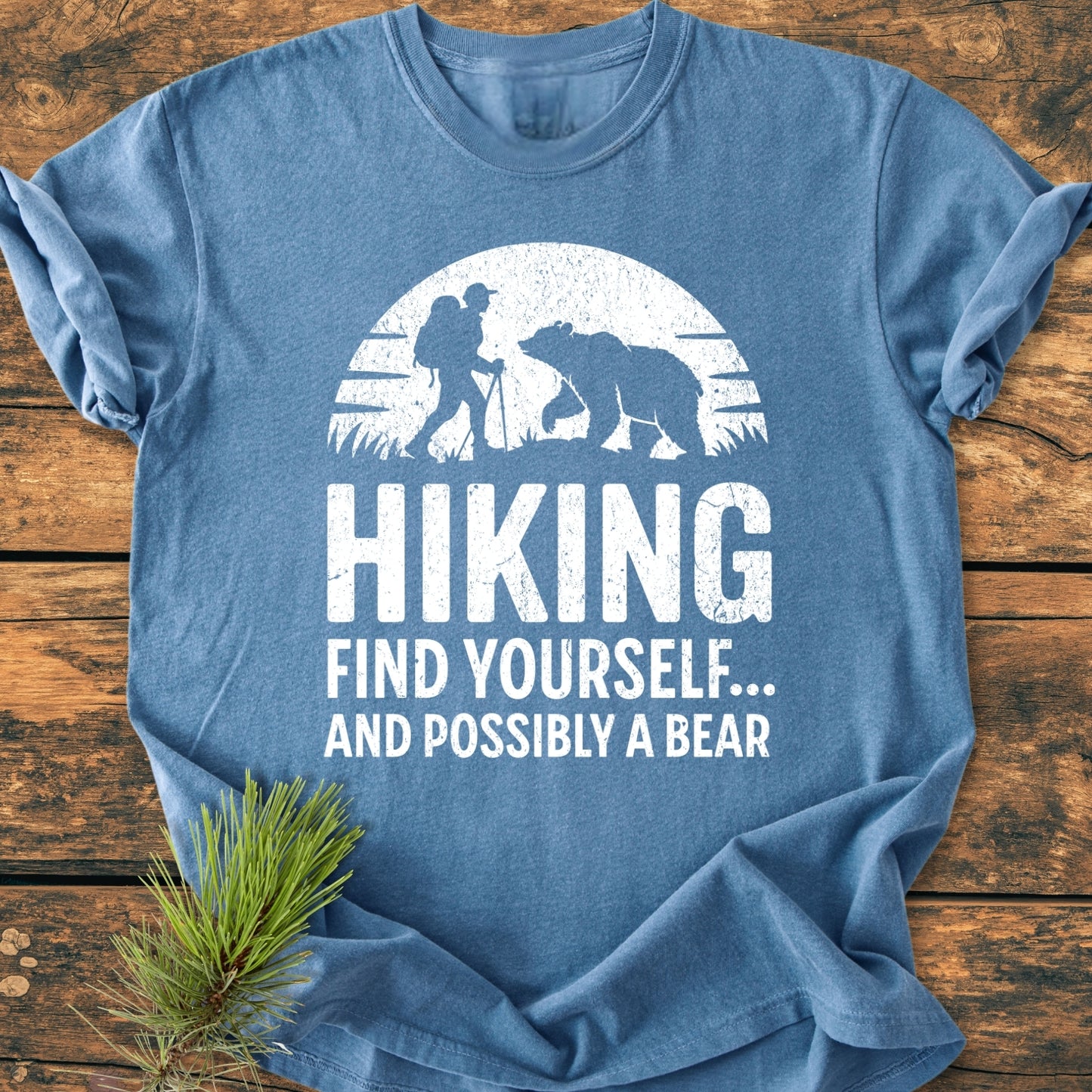 Find Yourself And Possibly A Bear - Vintage Wash Tee features a graphic of a hiker and bear silhouette on soft heather-blue fabric, styled with a pine branch on a wooden surface.