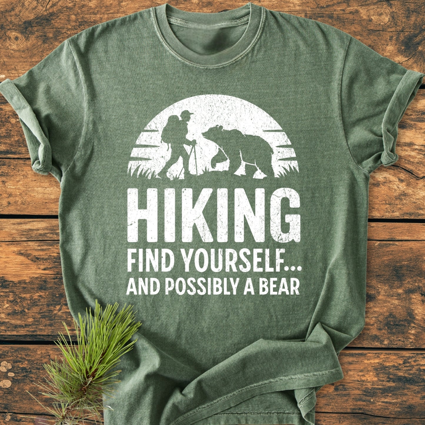 Find Yourself And Possibly A Bear - Vintage Wash Tee featuring a hiker versus bear graphic on forest-green, laid on rustic wood with pine sprig. Made from soft, durable, 100% ring-spun cotton.