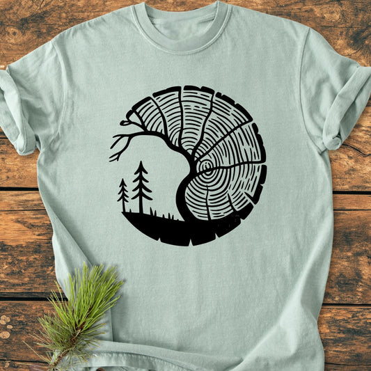 Tree Lover - Vintage Wash Tee displays a bold black yin-yang emblem with tree rings and pine silhouette on a pale green/gray surface, conveying nature's harmony. Beside it, a pine sprig enhances the theme.