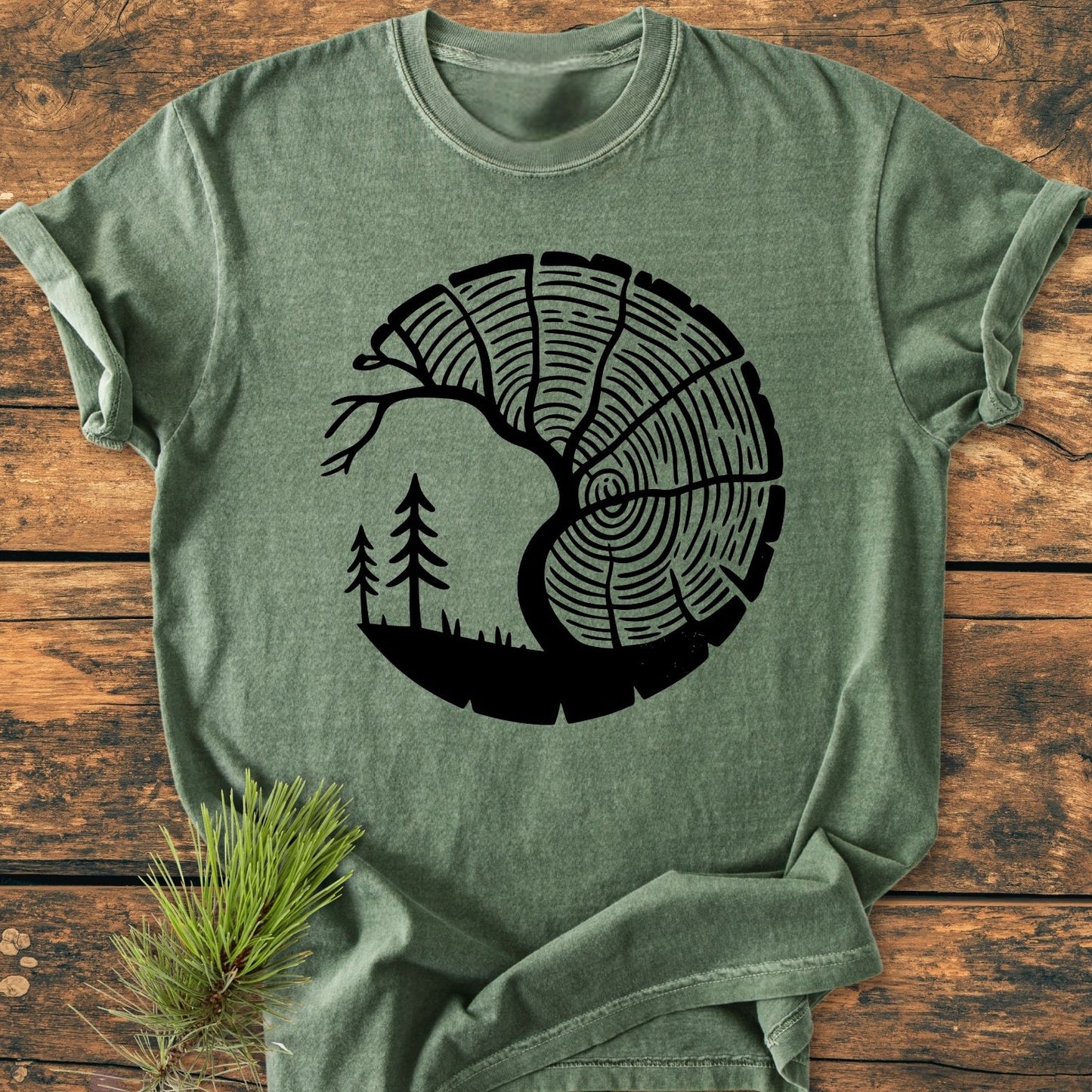 Tree Lover - Vintage Wash Tee features a unique half-tree-ring motif merging forest scenery on a heather-green T-shirt, laid on rustic wood. Pine sprig adds a woodland touch.
