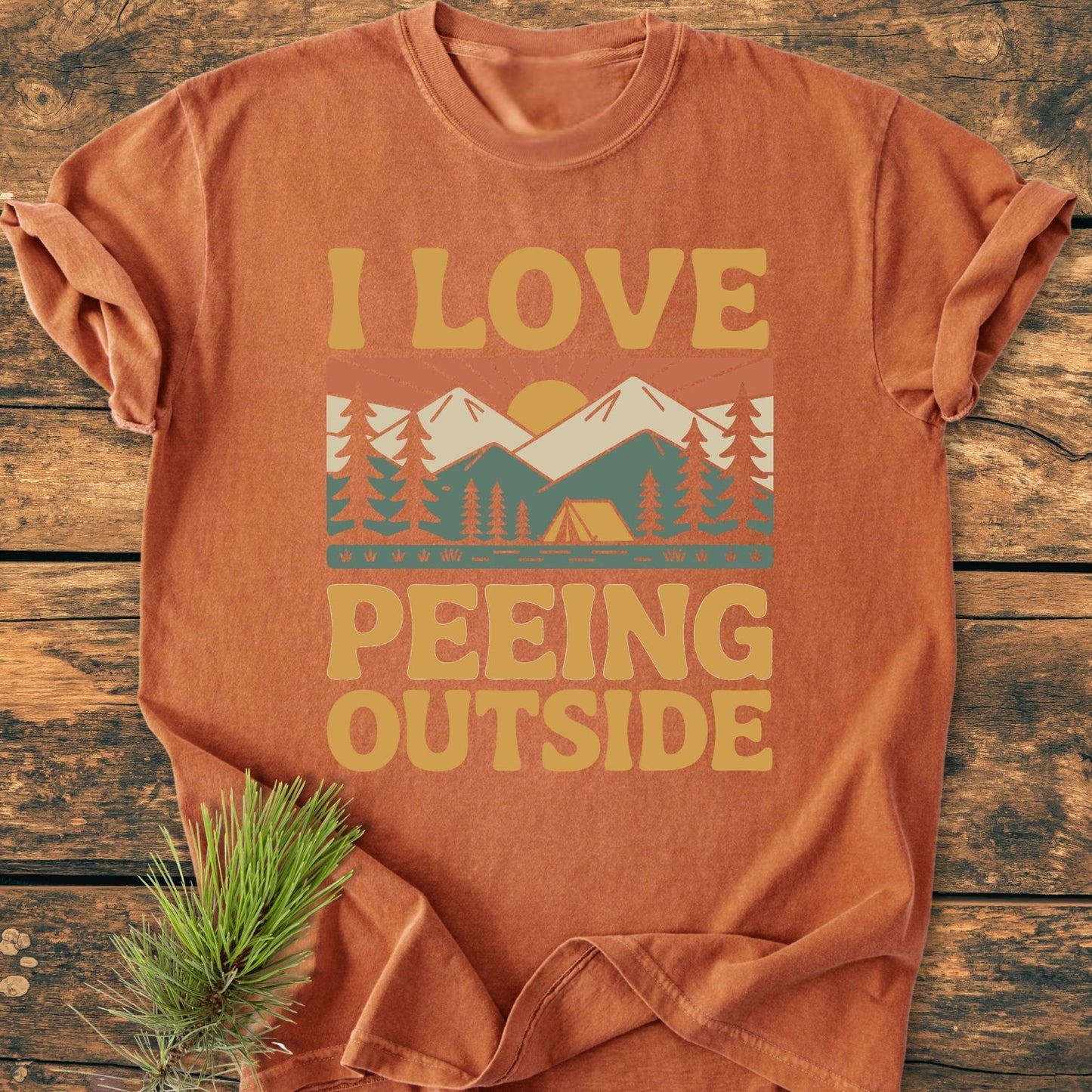I Love Peeing Outside - Vintage Wash Tee, displaying a stylized outdoor scene with pine trees and a tent, folded on wood with a pine sprig, highlights its rustic camping theme.