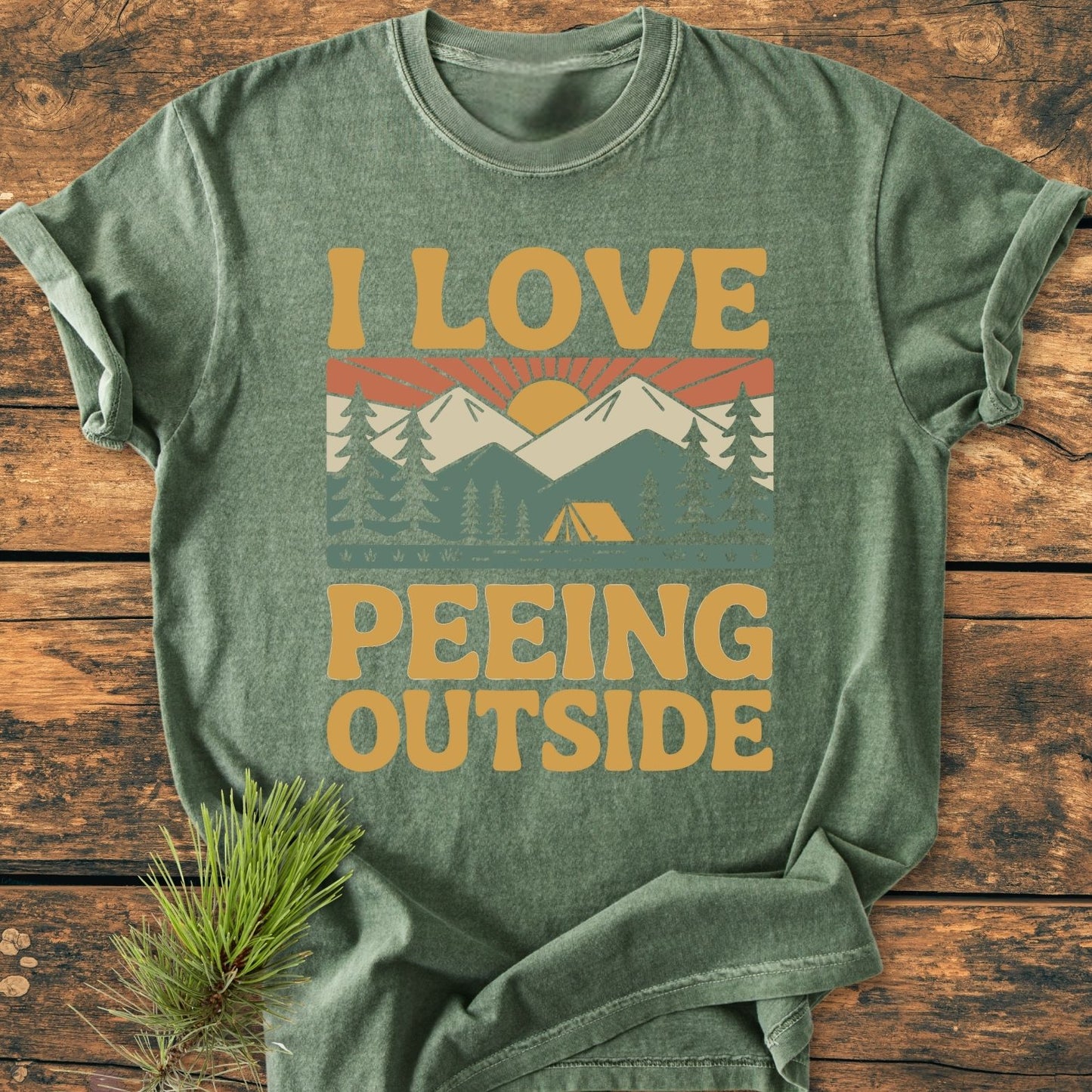 I Love Peeing Outside - Vintage Wash Tee features a retro outdoor graphic with pine trees, mountains, sun, and tent on a green heathered T-shirt, styled with a pine sprig beside it.