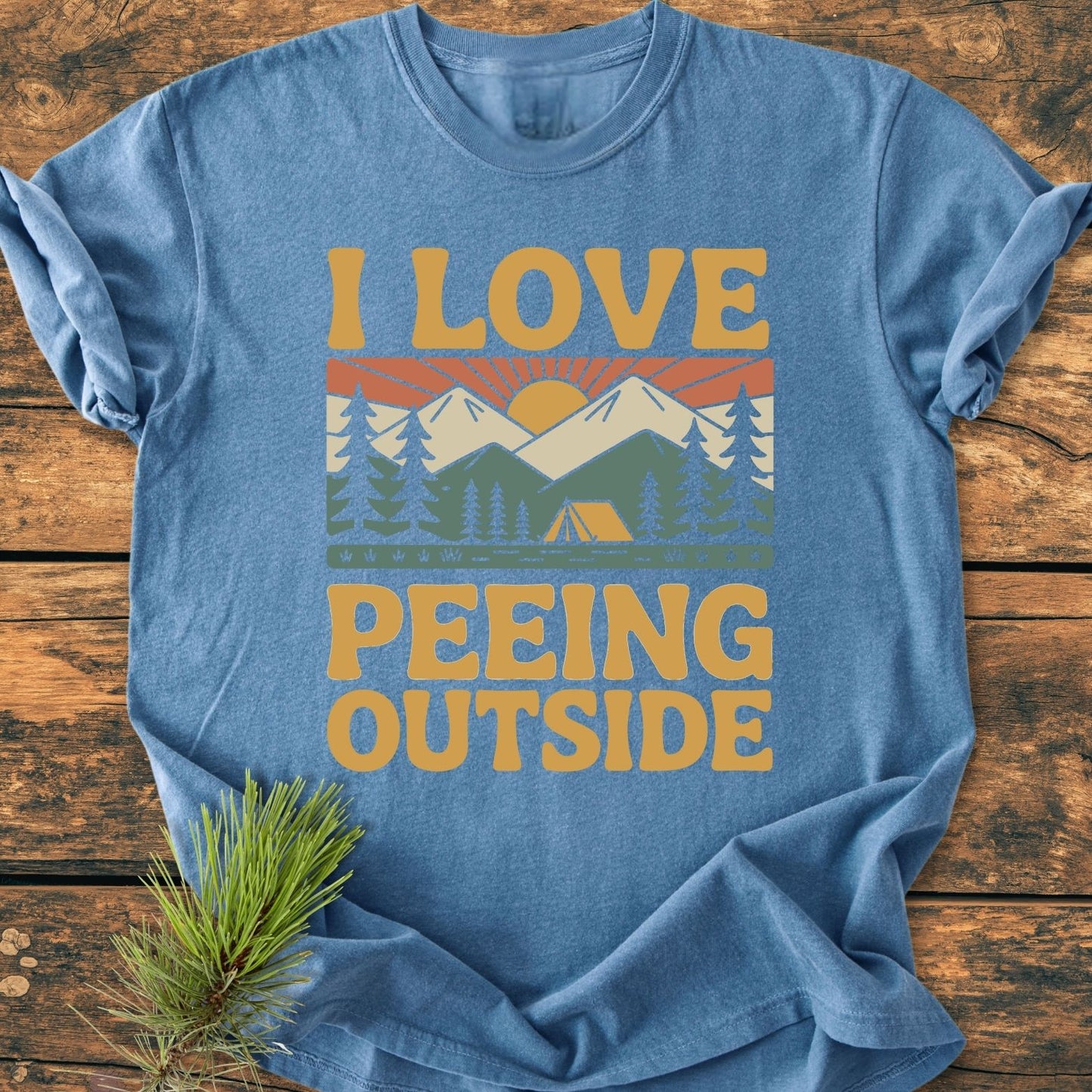 I Love Peeing Outside - Vintage Wash Tee features a screen-printed outdoor scene with mountains and trees, on a heather-blue, relaxed-fit, ring-spun cotton shirt, laid on a wooden background.