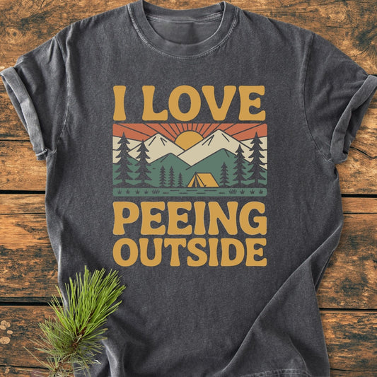 Charcoal-gray I Love Peeing Outside - Vintage Wash Tee featuring a retro graphic with pine trees, tent, mountains, and rising sun, on a wooden surface. Comfortable, 100% ring-spun cotton with a relaxed fit.