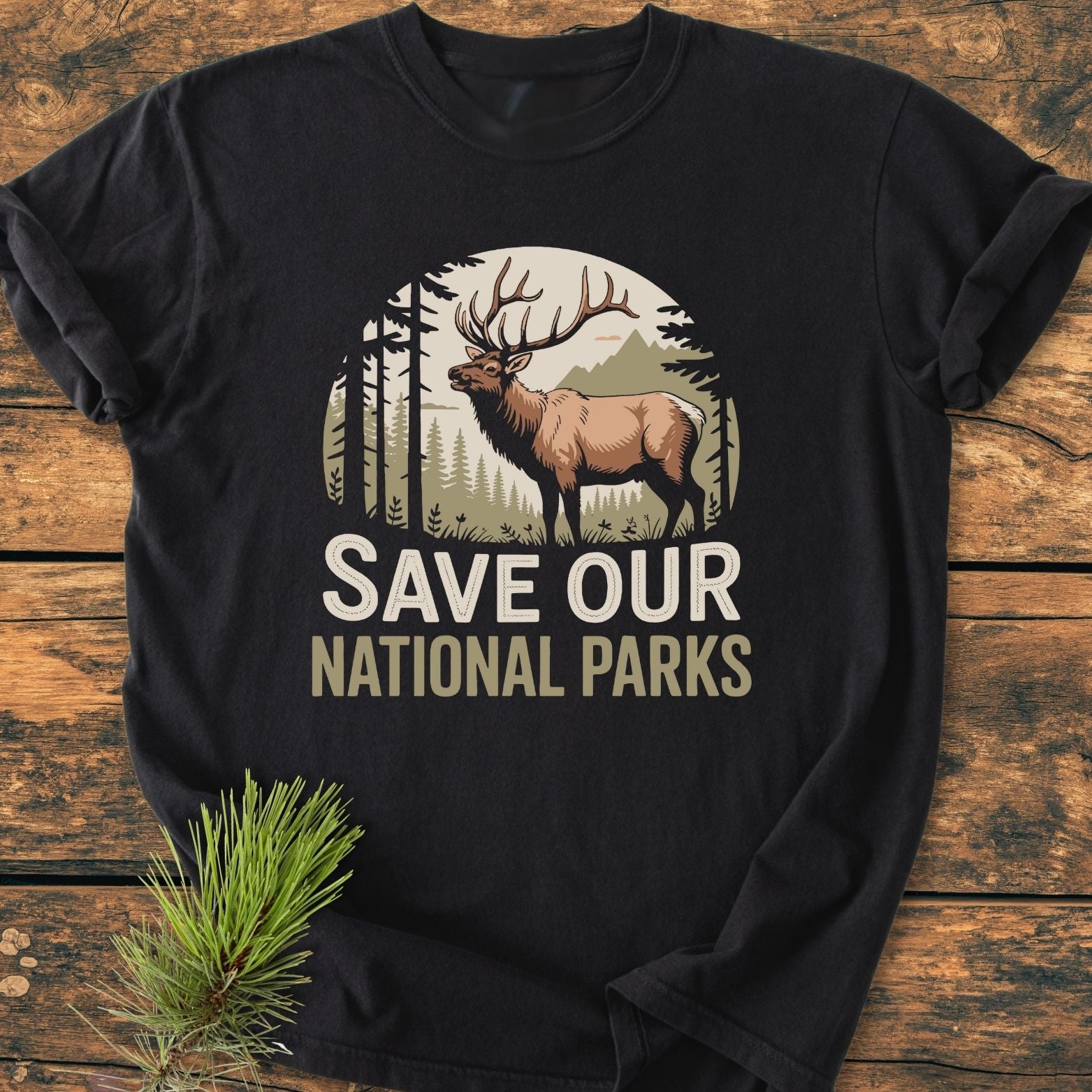 Save Our National Parks - Vintage Wash Tee features a nature-themed elk graphic against trees and mountains, on a black garment-dyed, soft-washed T-shirt, laid on wood with a pine sprig.