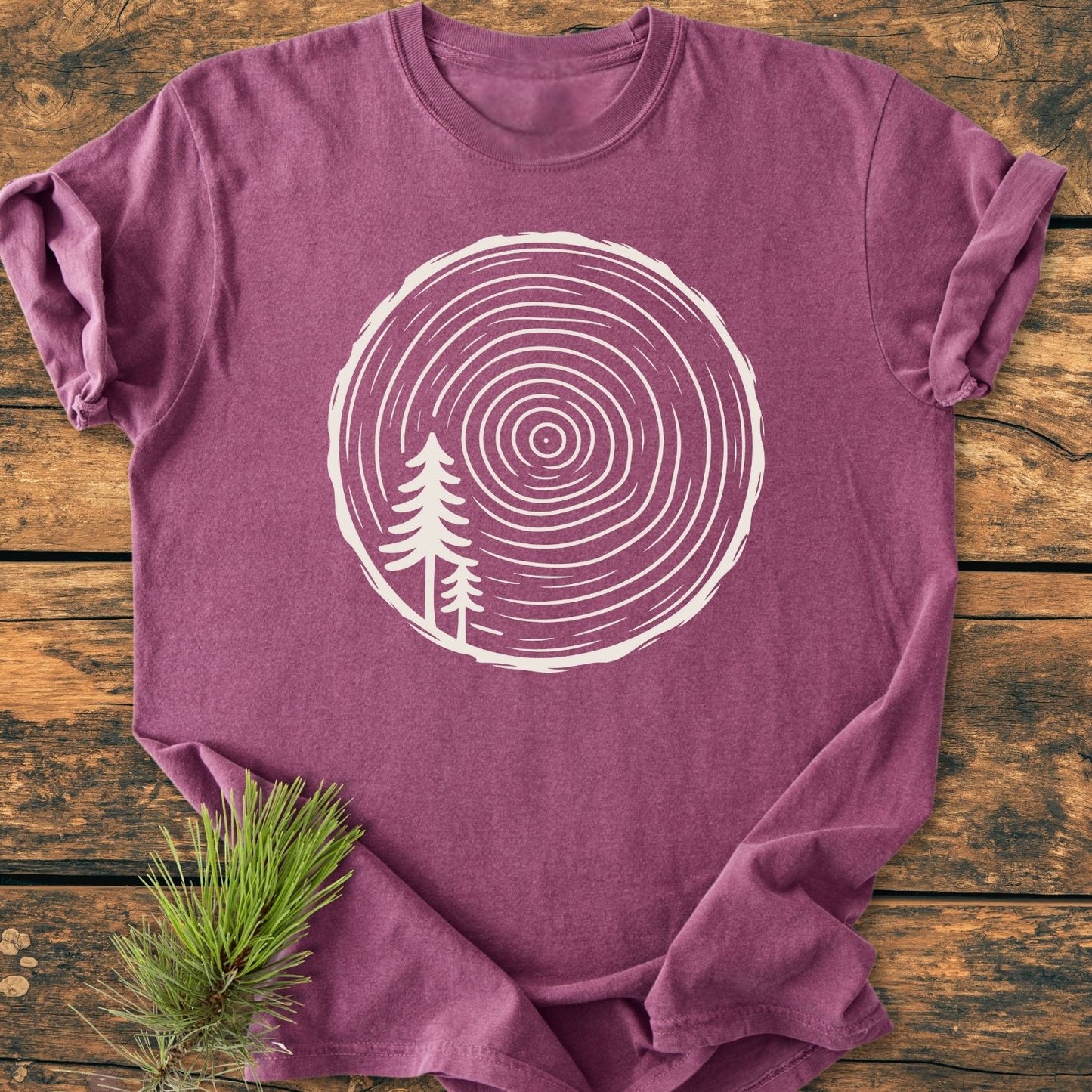 Minimalist Log - Vintage Wash Tee featuring a white tree ring graphic with pine silhouettes on a plum crew-neck, laid on rustic wood. Made of soft, durable garment-dyed cotton for comfort.
