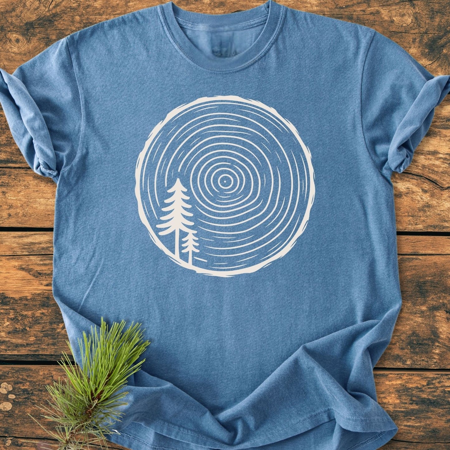 Minimalist Log - Vintage Wash Tee in heather-blue, featuring a white tree-ring graphic and stylized pine trees. Laid on wooden surface, highlighting its relaxed fit and durable construction with nearby pine sprig.