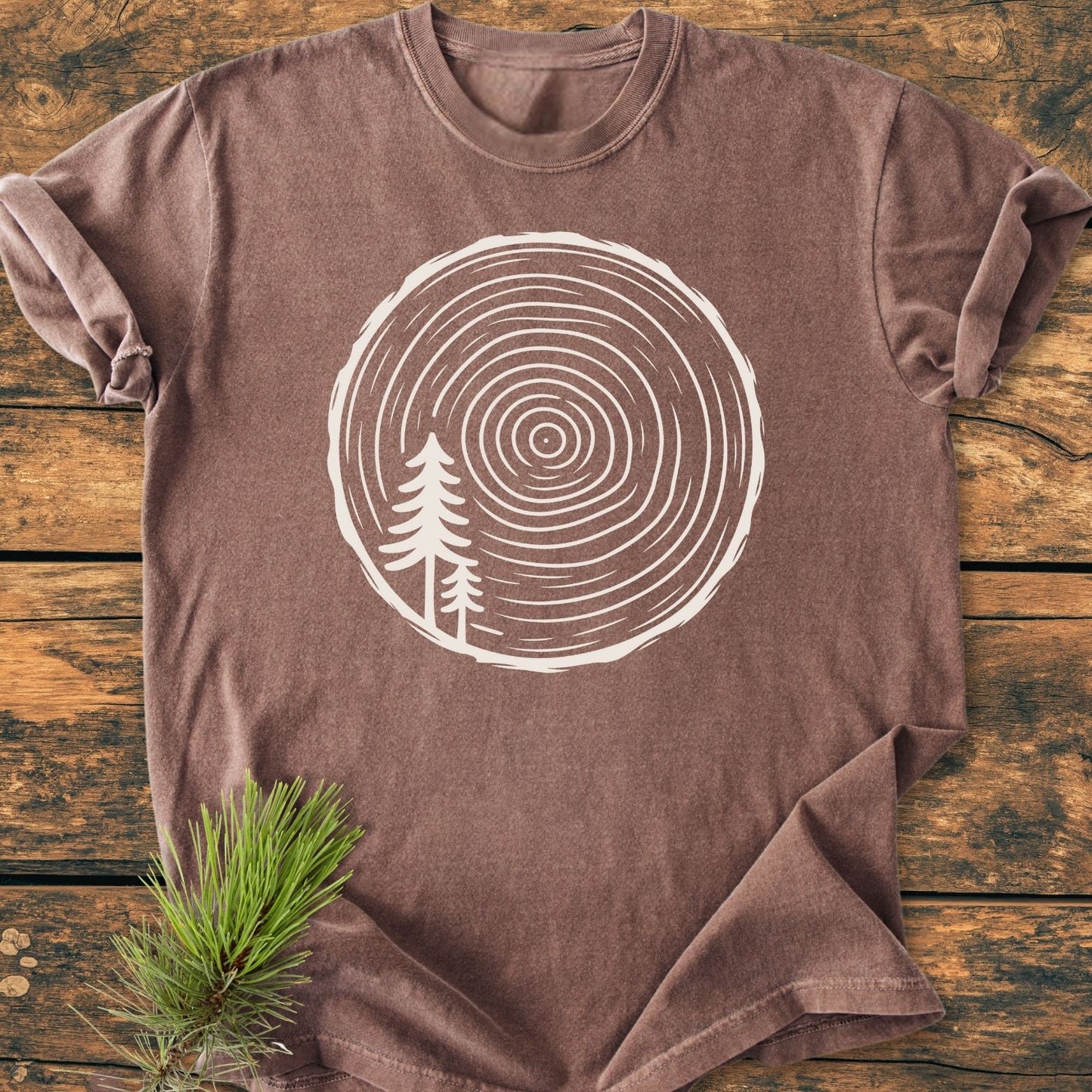 Minimalist Log - Vintage Wash Tee: Brown short-sleeve T-shirt featuring a white tree rings graphic with pine silhouettes, laid on wood. Nearby, a small pine branch complements the rustic design.