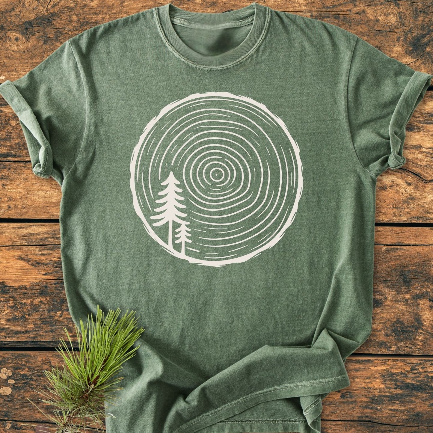 Minimalist Log - Vintage Wash Tee in forest-green with a white tree-ring graphic and evergreen silhouettes, laid on a wooden surface with a pine sprig. Crew-neck, relaxed fit, 100% ring-spun cotton.