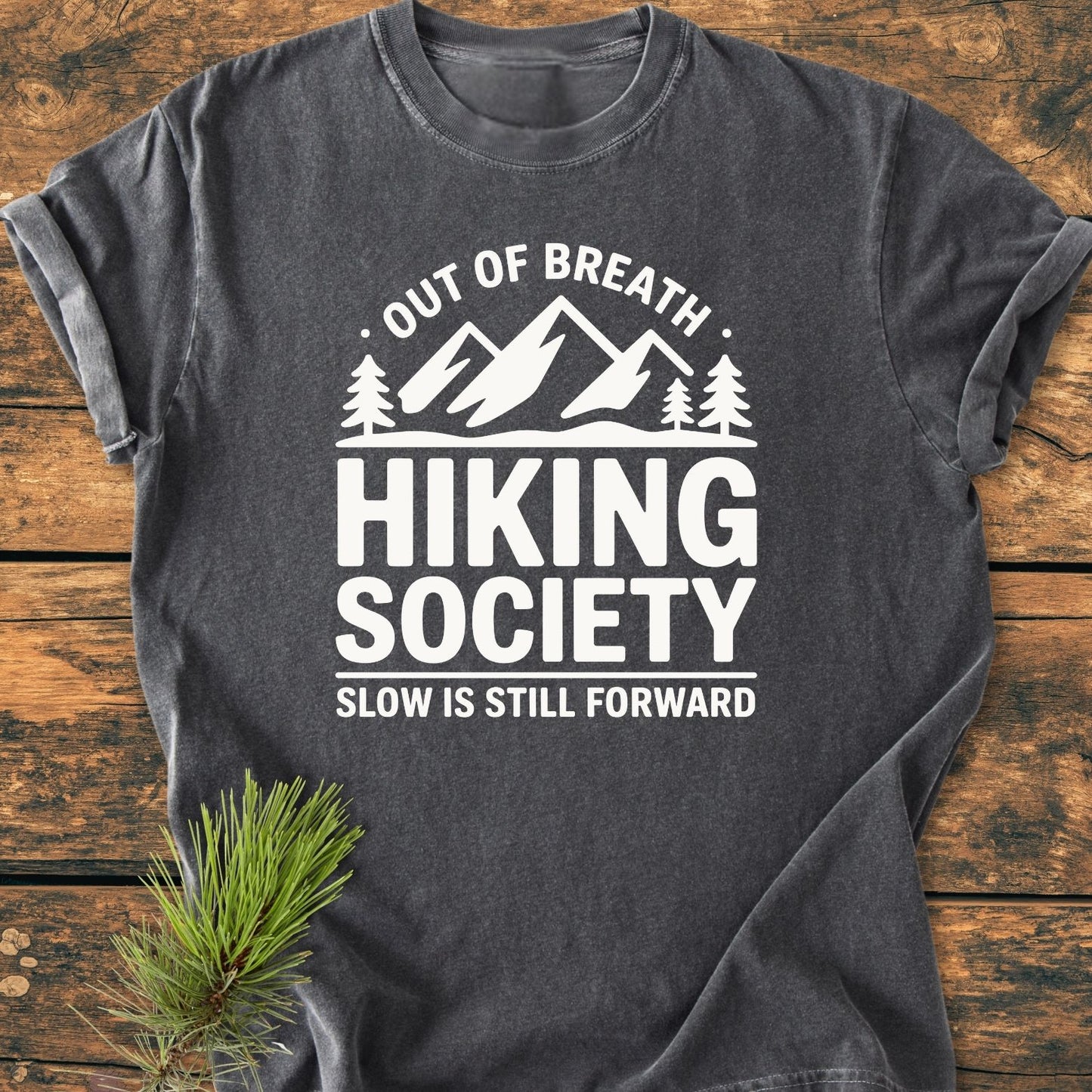 Out Of Breath - Vintage Wash Tee: Charcoal-gray T-shirt on a wooden surface, featuring a white mountain-and-trees logo with text, showcasing its relaxed fit and soft-washed, garment-dyed fabric.