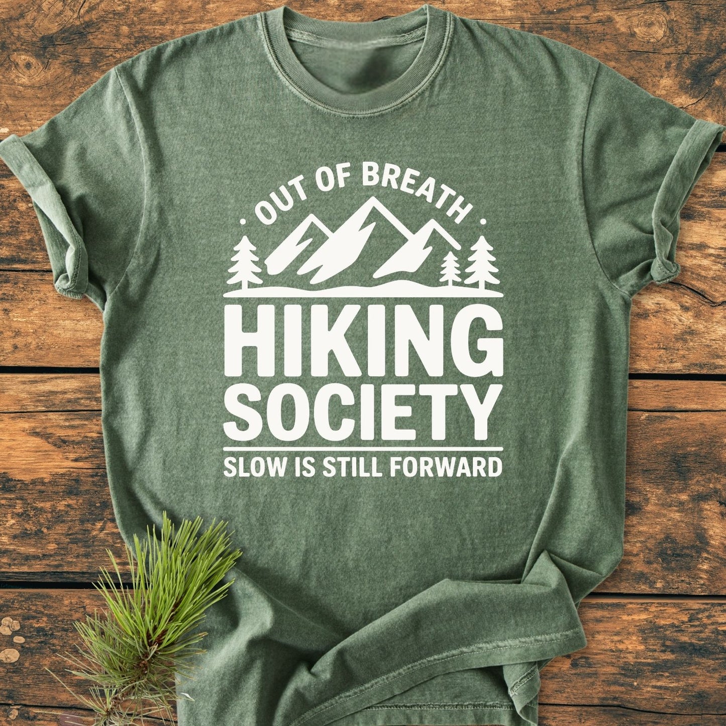 Out Of Breath - Vintage Wash Tee featuring a stylized mountain and pine design, laid flat on wood. The shirt emphasizes outdoor adventure with slogans and offers a cozy, relaxed fit.