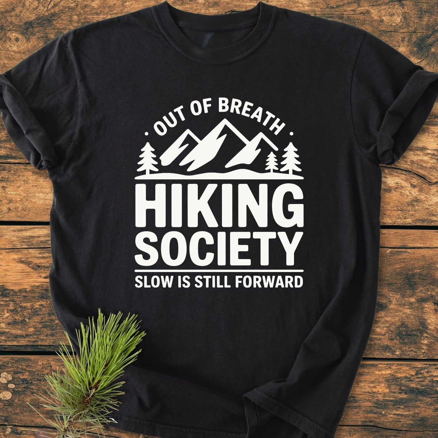 Out Of Breath - Vintage Wash Tee featuring a mountain-and-trees graphic with hiking-themed text, displayed on rustic wood with a pine branch, highlighting its relaxed, durable design made from soft, garment-dyed cotton.