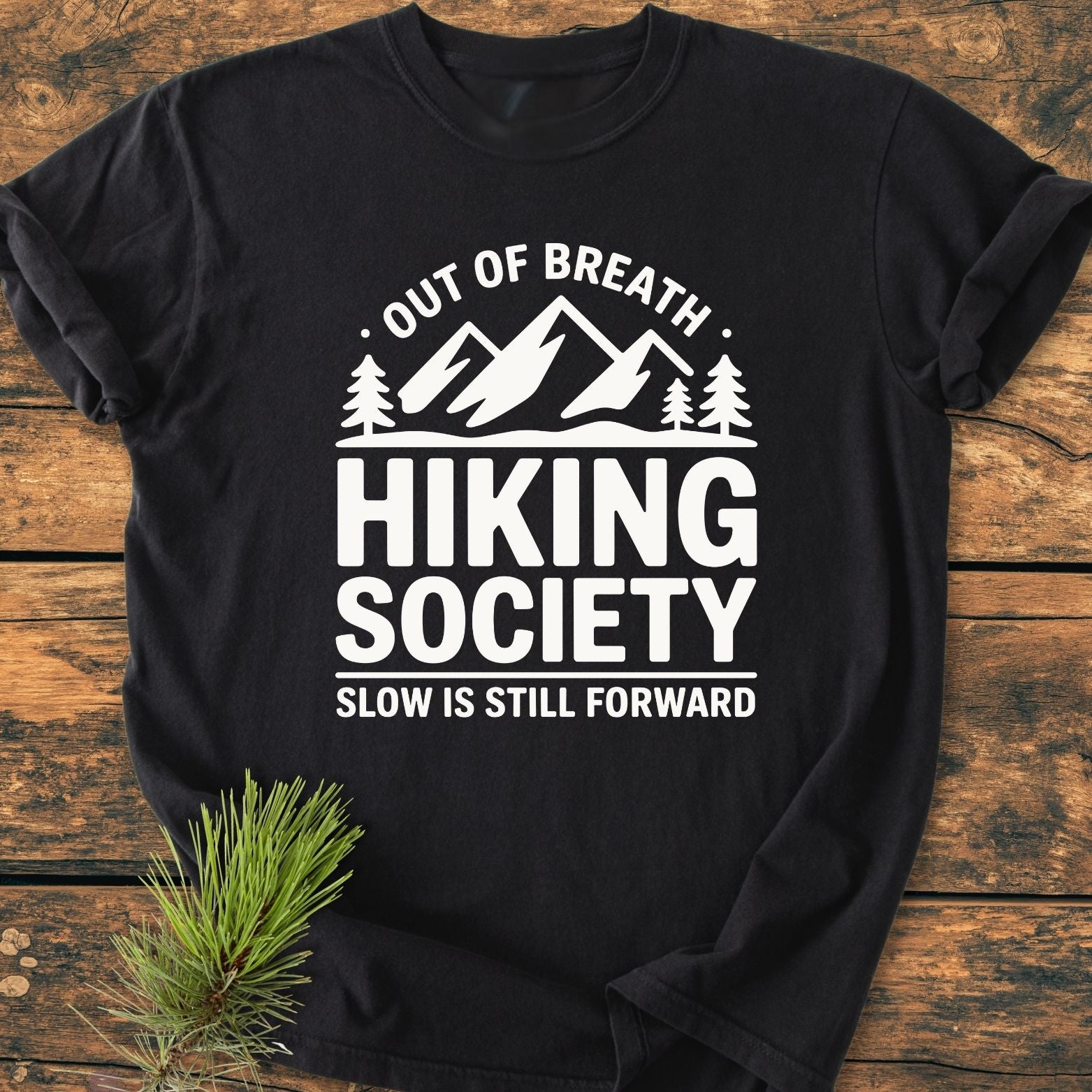 Out Of Breath - Vintage Wash Tee featuring a mountain-and-trees graphic with hiking-themed text, displayed on rustic wood with a pine branch, highlighting its relaxed, durable design made from soft, garment-dyed cotton.