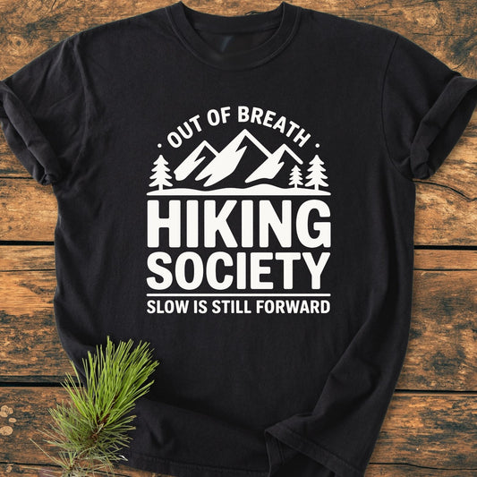 Out Of Breath - Vintage Wash Tee featuring a mountain-and-trees graphic with hiking-themed text, displayed on rustic wood with a pine branch, highlighting its relaxed, durable design made from soft, garment-dyed cotton.