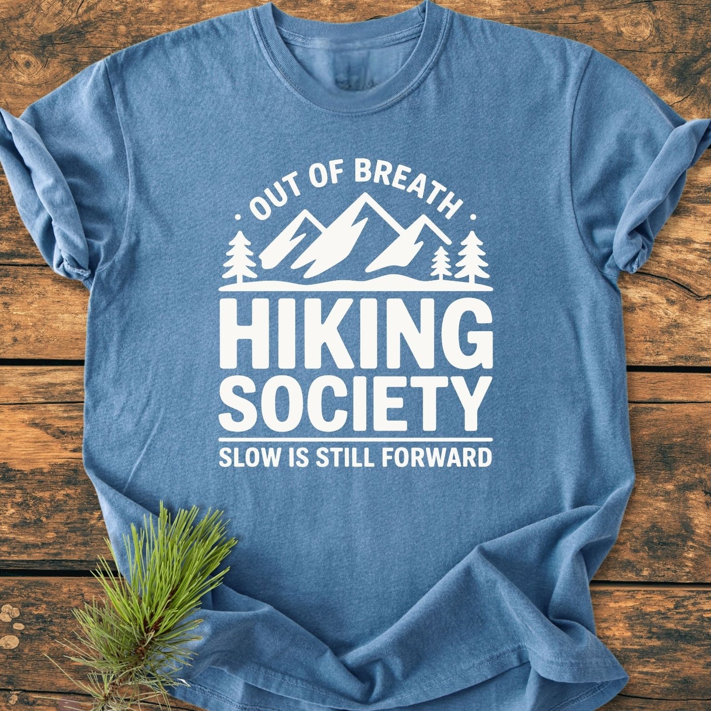Out Of Breath - Vintage Wash Tee features a mountain and pine tree print with “Hiking Society” and “Slow is Still Forward” slogans, displayed on a heathered blue crew-neck T-shirt.