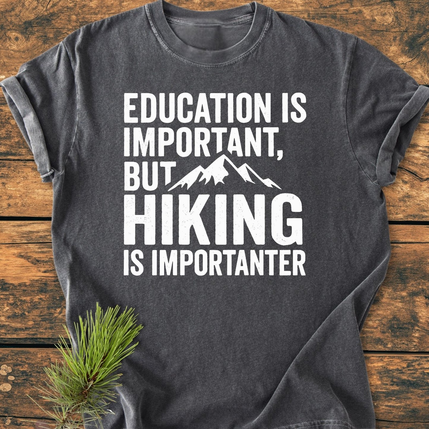 Education Is Important - Vintage Wash Tee displayed on a wooden surface, features a humorous hiking slogan with a mountain graphic. Soft-washed cotton offers comfort, complemented by durable stitching and a relaxed fit.