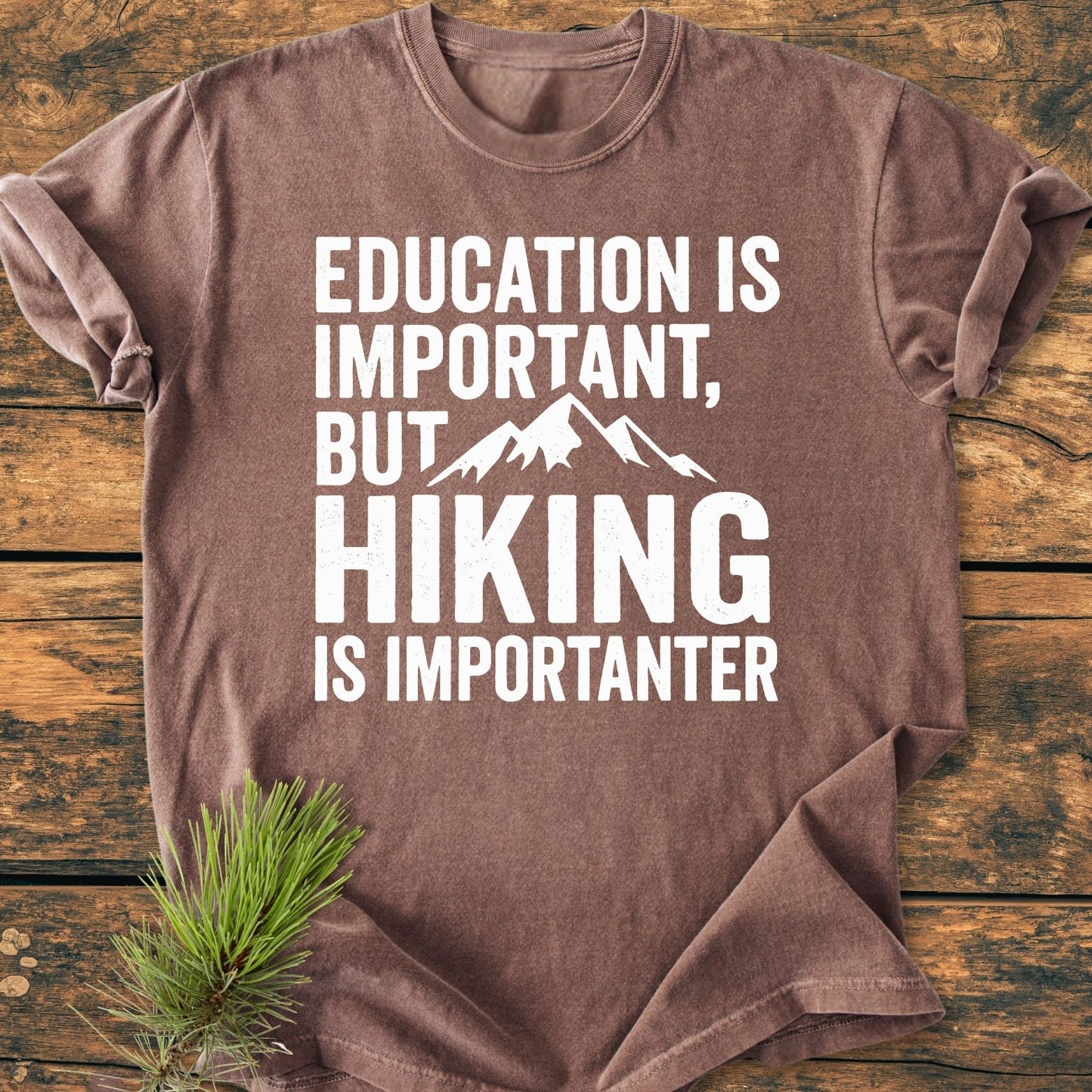 Education Is Important - Vintage Wash Tee features a brown crew-neck with a mountain-themed slogan. Displayed on wood, it includes a pine sprig accent, showcasing a cozy, relaxed fit and durable construction.