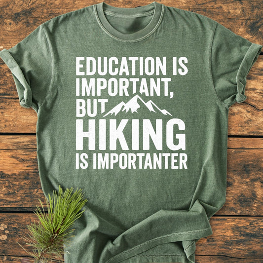 Education Is Important - Vintage Wash Tee in heather-green features a playful slogan and mountain silhouette, laid flat on a wooden surface with pine needles nearby, showcasing its cozy, relaxed fit.