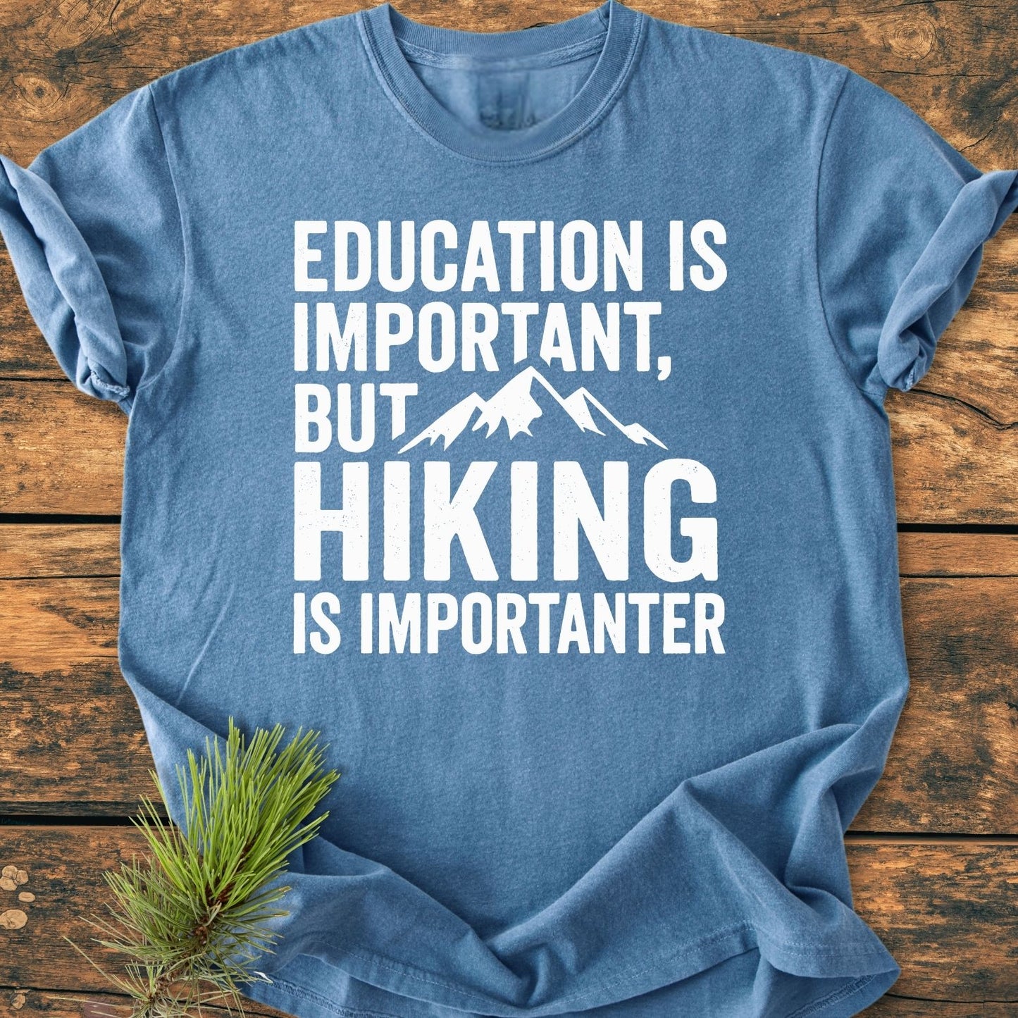 Heather-blue Education Is Important - Vintage Wash Tee with bold white lettering and a mountain graphic, laid flat on wood. Pine needles accent the soft, garment-dyed, relaxed-fit, ring-spun cotton shirt.