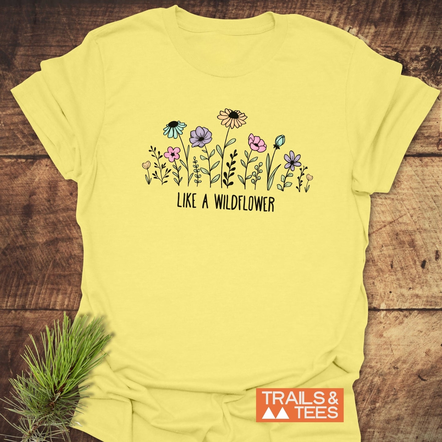 Like A Wildflower T-Shirt featuring hand-drawn spring wildflowers and the phrase “Like a Wildflower” in black, set against a rustic wooden backdrop, capturing an outdoorsy essence with pine branches.