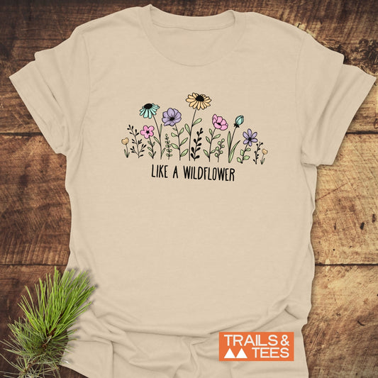 Like A Wildflower T-Shirt featuring hand-drawn wildflowers and the phrase “LIKE A WILDFLOWER,” with a durable print and TRAILS & TEES logo, displayed on a wooden surface.