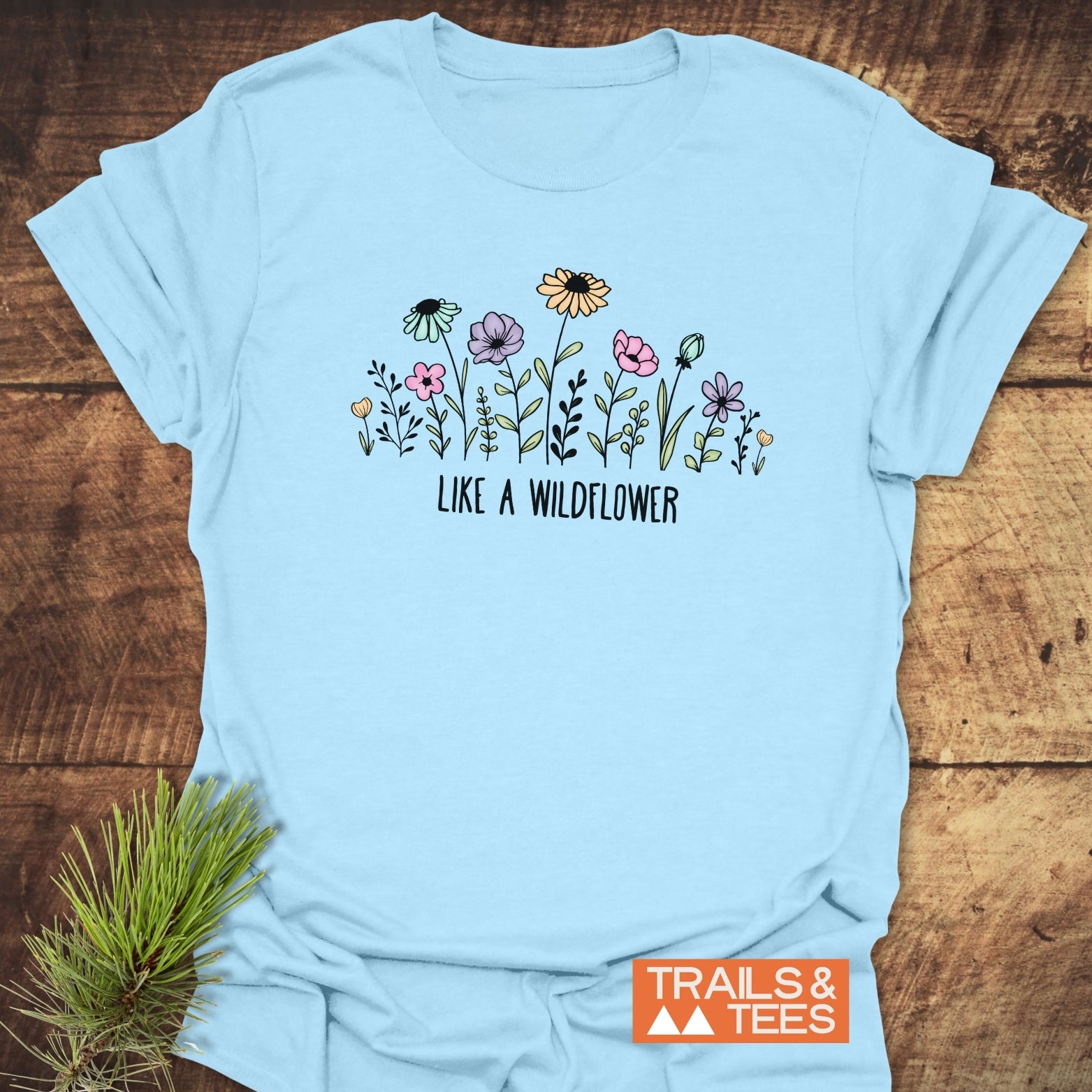 Like A Wildflower T-Shirt featuring hand-drawn wildflowers and 'LIKE A WILDFLOWER' slogan on chest, with an orange 'TRAILS & TEES' logo, laying on a wooden surface.