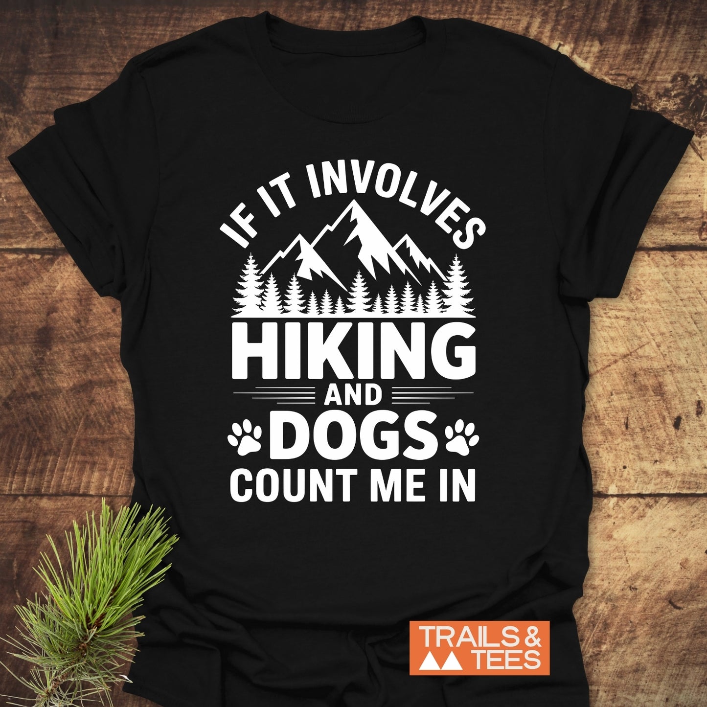 Hiking And Dogs T-Shirt featuring a white mountain-and-forest graphic with the text IF IT INVOLVES HIKING AND DOGS COUNT ME IN and paw-print icons, laid on a wooden surface.