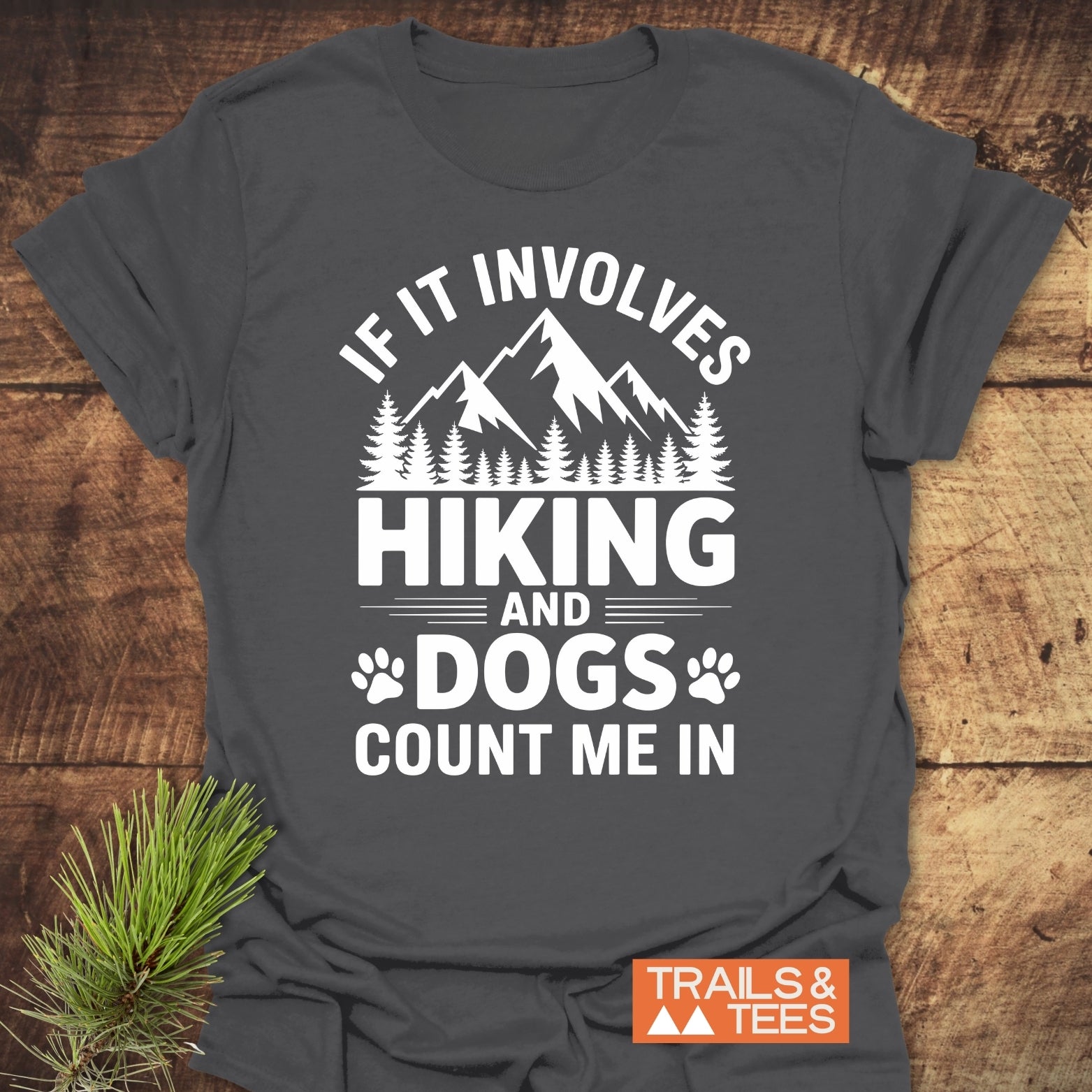 Hiking And Dogs T-Shirt featuring a white mountain and tree silhouette with paw prints and slogan on dark fabric. Includes Trails & Tees logo, symbolizing outdoor adventure and love for hiking with dogs.