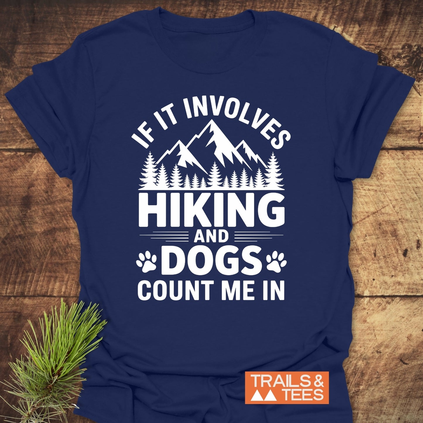Navy-blue Hiking And Dogs T-Shirt featuring a white mountain and forest graphic with paw prints, plus the slogan “IF IT INVOLVES HIKING AND DOGS COUNT ME IN,” on a wooden surface.