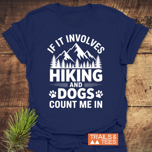 Navy-blue Hiking And Dogs T-Shirt featuring a white mountain and forest graphic with paw prints, plus the slogan “IF IT INVOLVES HIKING AND DOGS COUNT ME IN,” on a wooden surface.