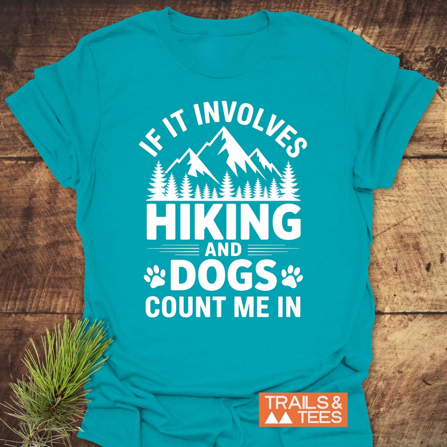 Teal Hiking And Dogs T-Shirt with white mountain-and-tree graphic and slogan, laid flat with pine sprig. Features TRAILS & TEES. logo, emphasizing outdoor adventure spirit.