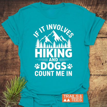 Teal Hiking And Dogs T-Shirt with white mountain-and-tree graphic and slogan, laid flat with pine sprig. Features TRAILS & TEES. logo, emphasizing outdoor adventure spirit.