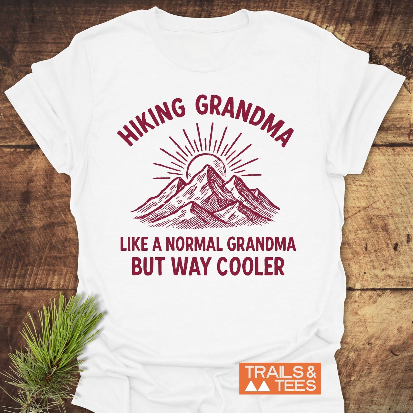 Cool Hiking Grandma Sunrise T-Shirt featuring a mountain-and-sunburst graphic with bold burgundy text, laid on a wooden surface with a pine sprig. Includes a small TRAILS & TEES logo.