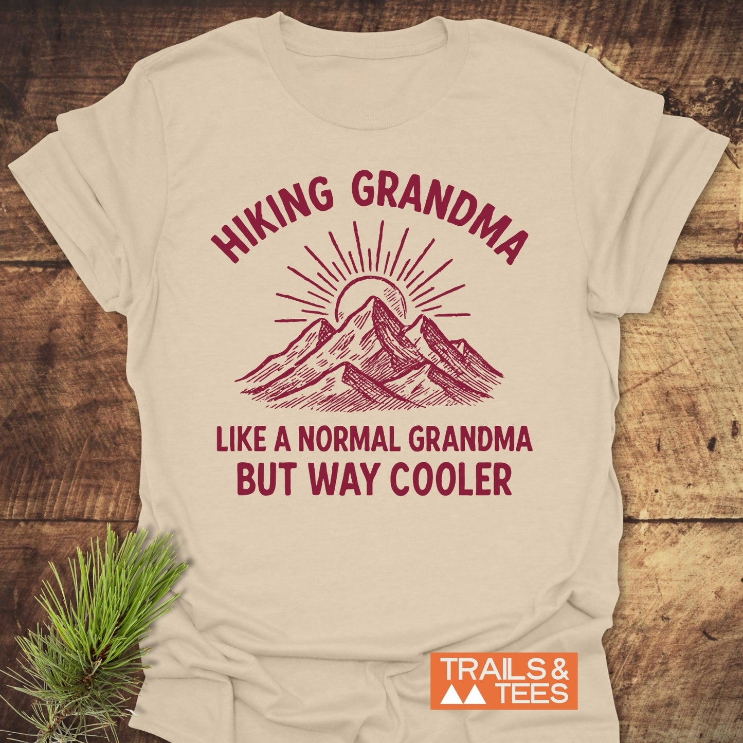 Cool Hiking Grandma Sunrise T-Shirt features a mountain and sun illustration with playful text, laid on wood with a pine sprig, branded by Trails & Tees logo.