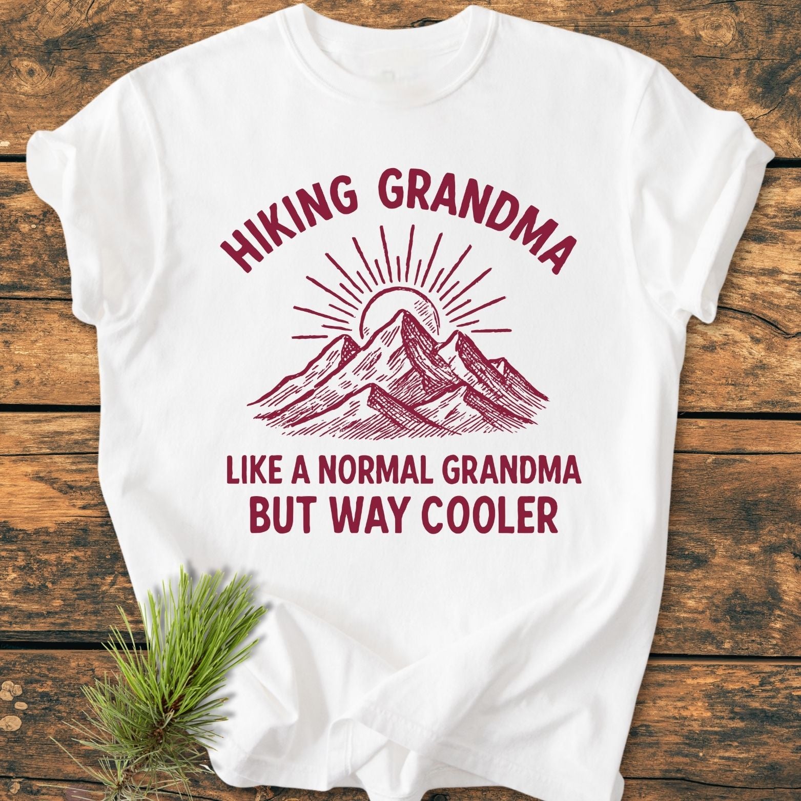 Cool Hiking Grandma Sunrise - Vintage Wash Tee on wood surface, featuring a red mountain and sunburst design with humorous text. A pine sprig accents the soft, durable, relaxed-fit shirt.