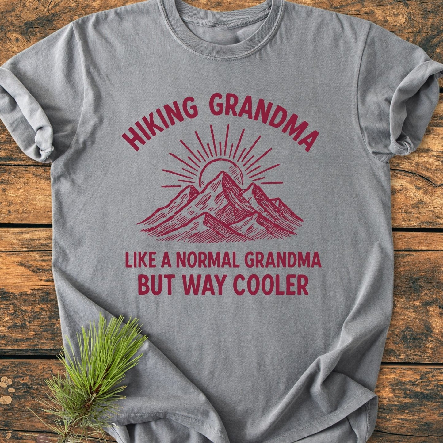 Cool Hiking Grandma Sunrise - Vintage Wash Tee, featuring a mountain and sun graphic in maroon on a gray background, laid flat on wood with pine needles beside. Comfortable, ring-spun cotton fabric.