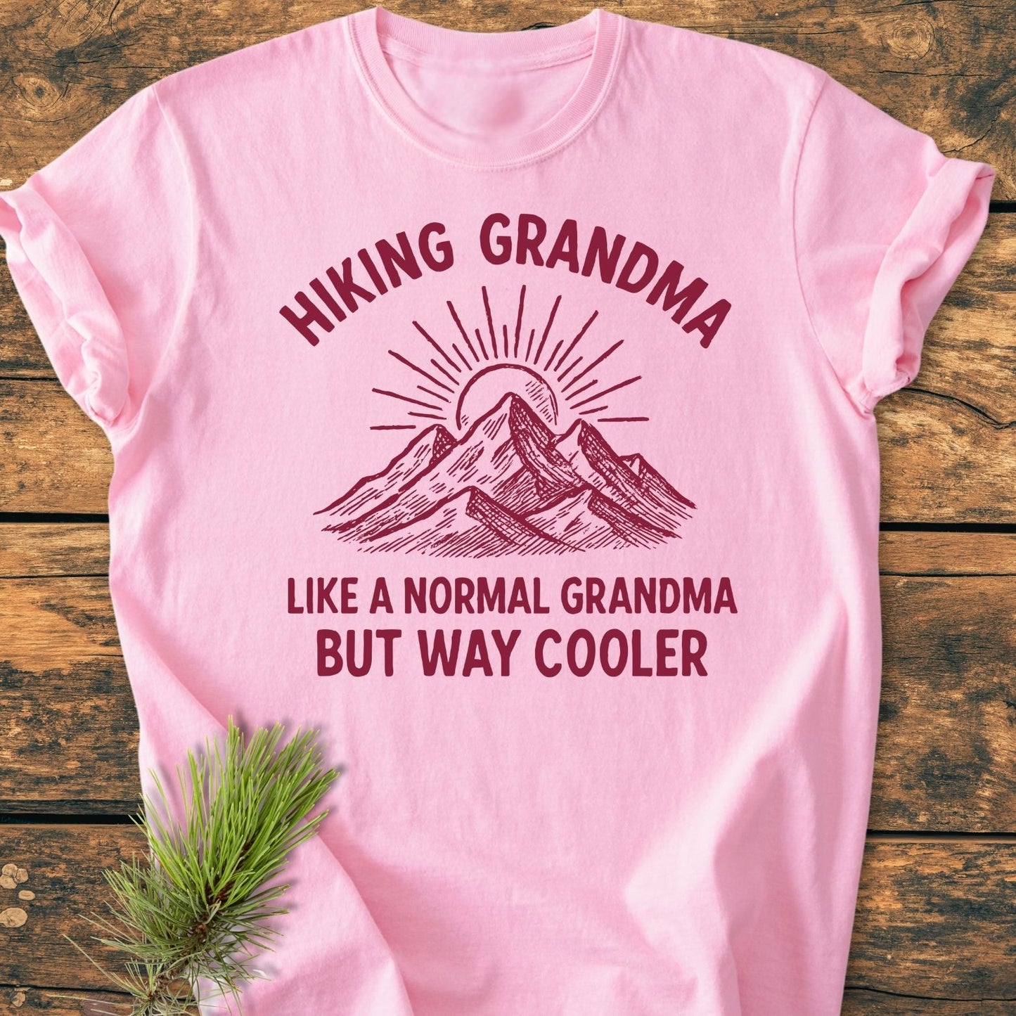 Cool Hiking Grandma Sunrise - Vintage Wash Tee features a dark-red mountain and sun graphic, slogan, on a light pink backdrop. Laid on wood-grain, embodies comfort with soft, durable ring-spun cotton.