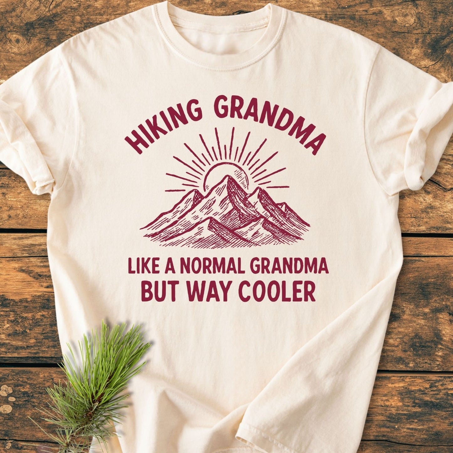 Cream-colored Cool Hiking Grandma Sunrise - Vintage Wash Tee featuring maroon text and mountain graphic with sun rays on rustic wood background, accented by a pine branch, highlighting its cozy, relaxed fit.