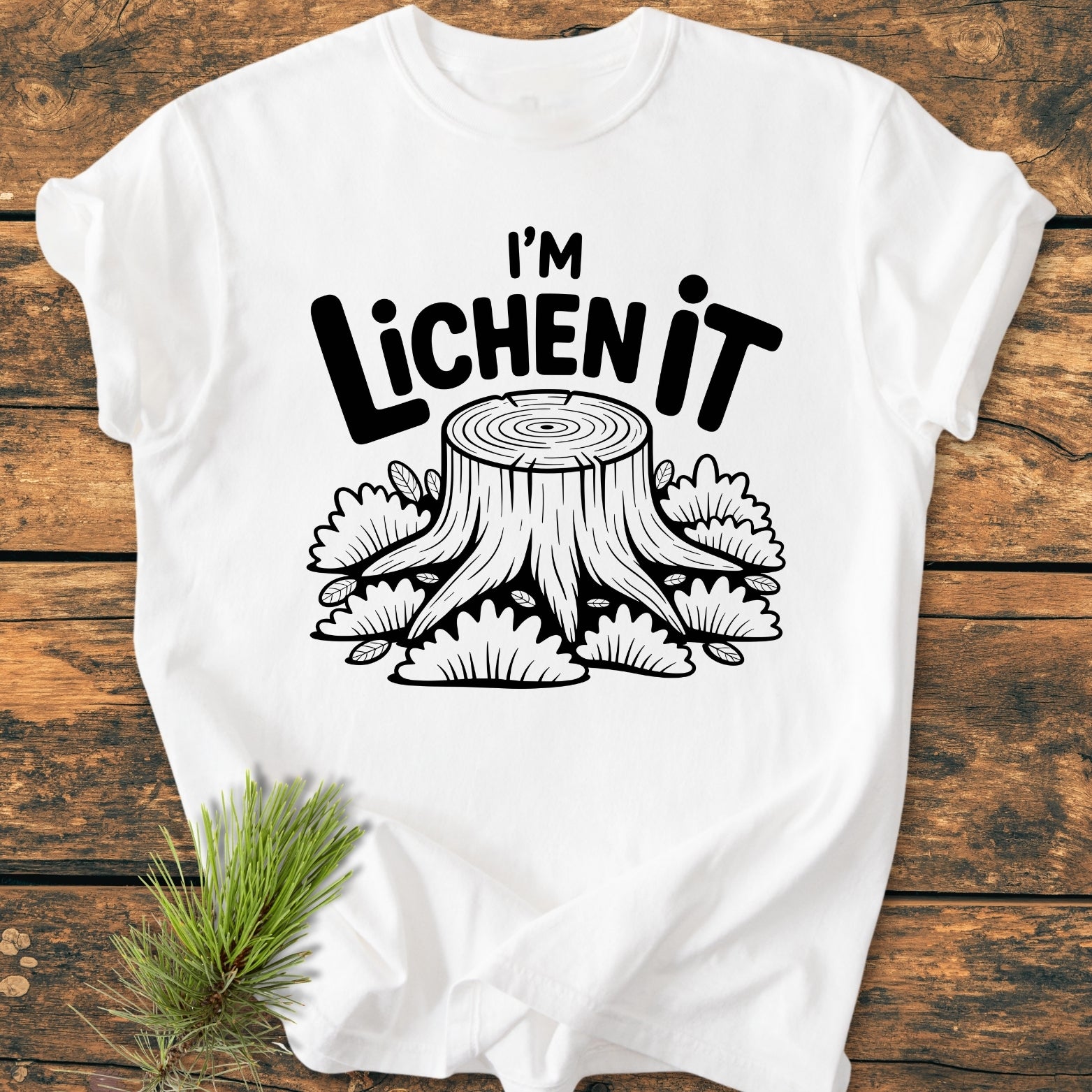 I'm Lichen It - Vintage Wash Tee featuring a cartoon tree-stump graphic with lichen, printed on a soft, white, garment-dyed t-shirt, laid on a rustic wooden surface with a pine sprig accent.