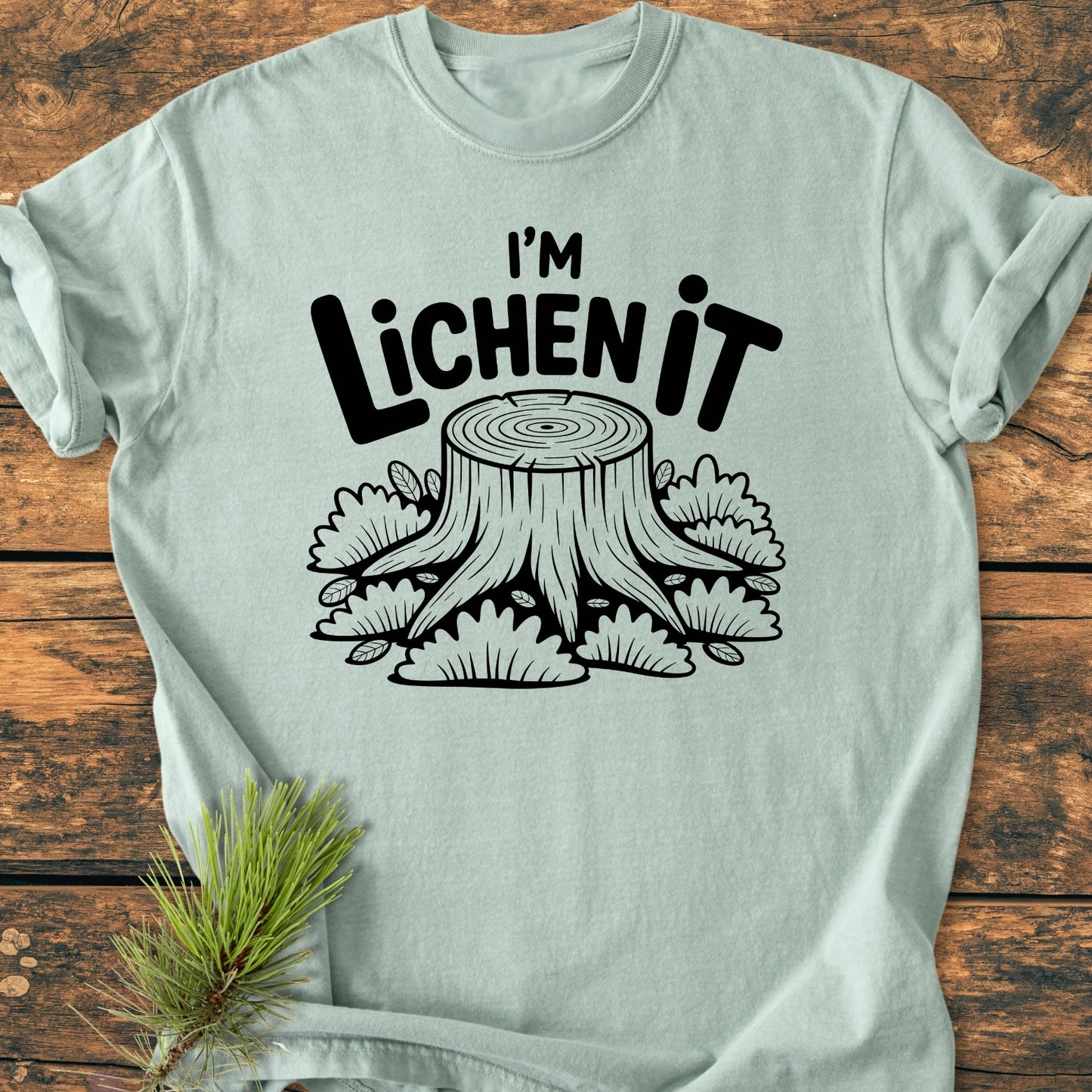 I'm Lichen It - Vintage Wash Tee featuring a stylized tree stump with lichen print on a light-green background, displayed on wood with a pine sprig for a nature-themed presentation.