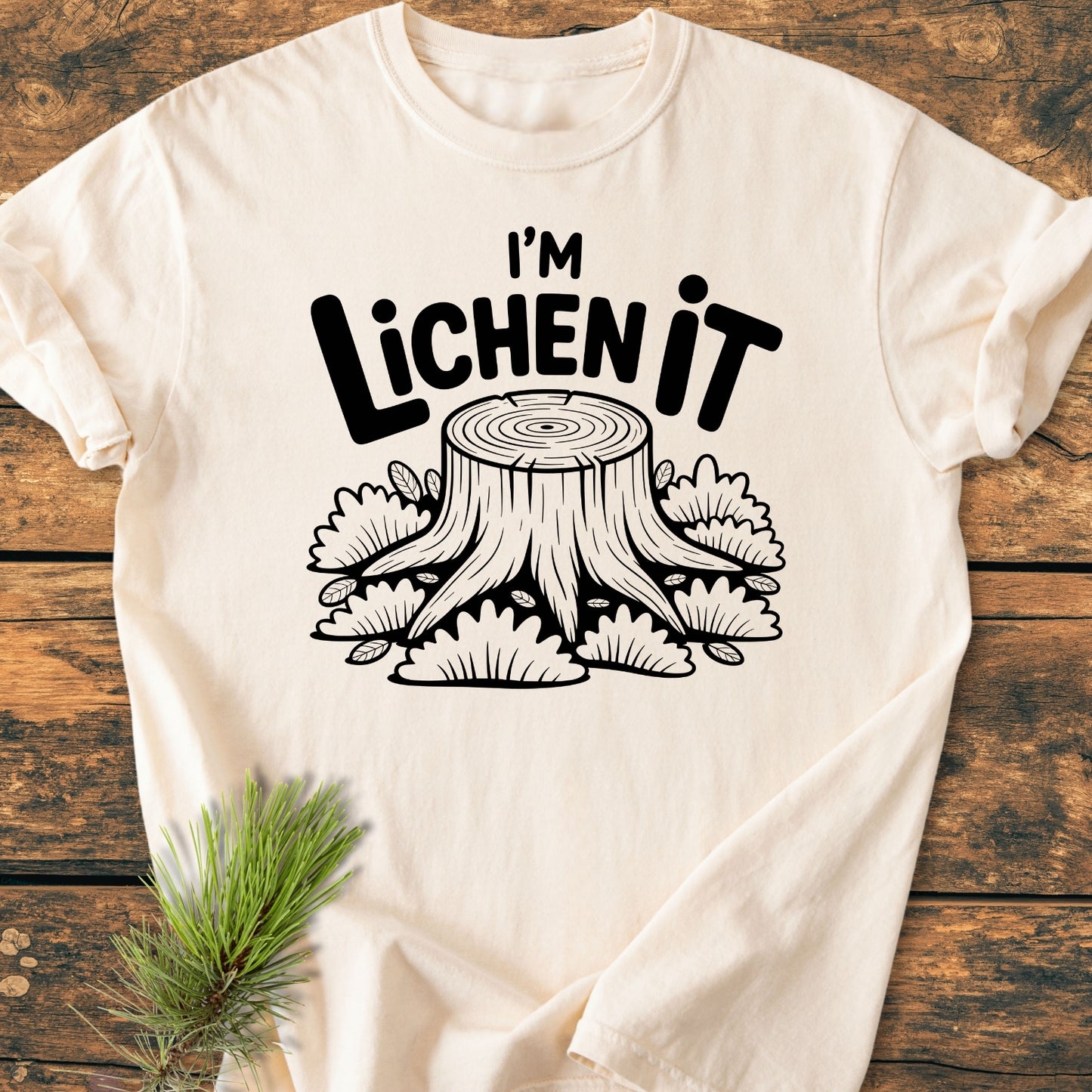 I'm Lichen It - Vintage Wash Tee featuring a cartoon tree stump and lichen with a pun slogan, laid on wood. Includes a pine sprig accent, highlighting its relaxed, durable design.