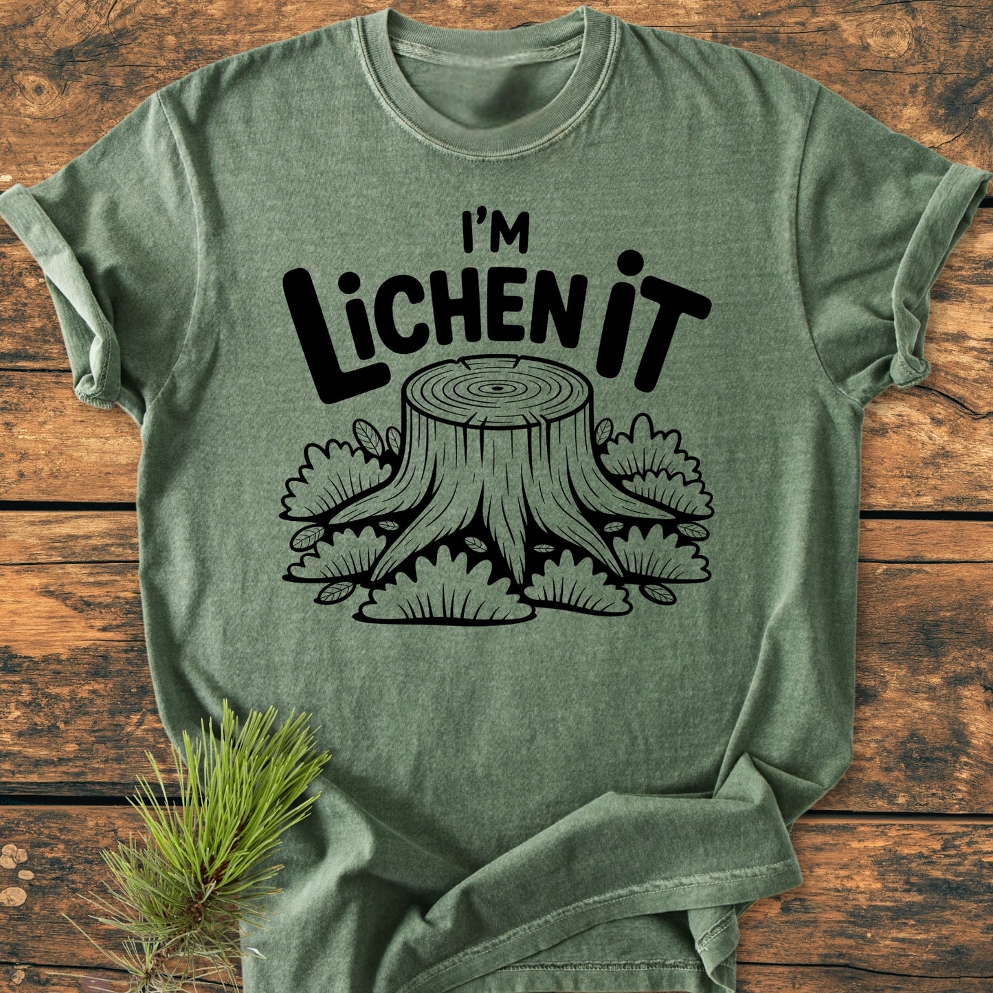 I'm Lichen It - Vintage Wash Tee features a black line-art tree stump design on a heather-green shirt, styled on rustic wood with a pine sprig, showcasing its relaxed fit and durability.