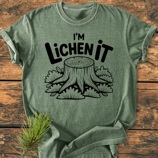 I'm Lichen It - Vintage Wash Tee features a black line-art tree stump design on a heather-green shirt, styled on rustic wood with a pine sprig, showcasing its relaxed fit and durability.