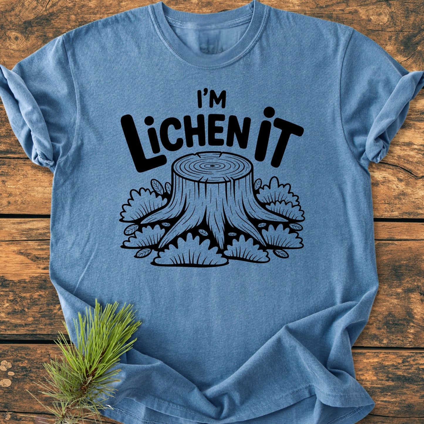 I'm Lichen It - Vintage Wash Tee features a heather-blue color with a black screen-printed tree stump and slogan, laid on wood with a pine sprig accent, showcasing its relaxed fit.